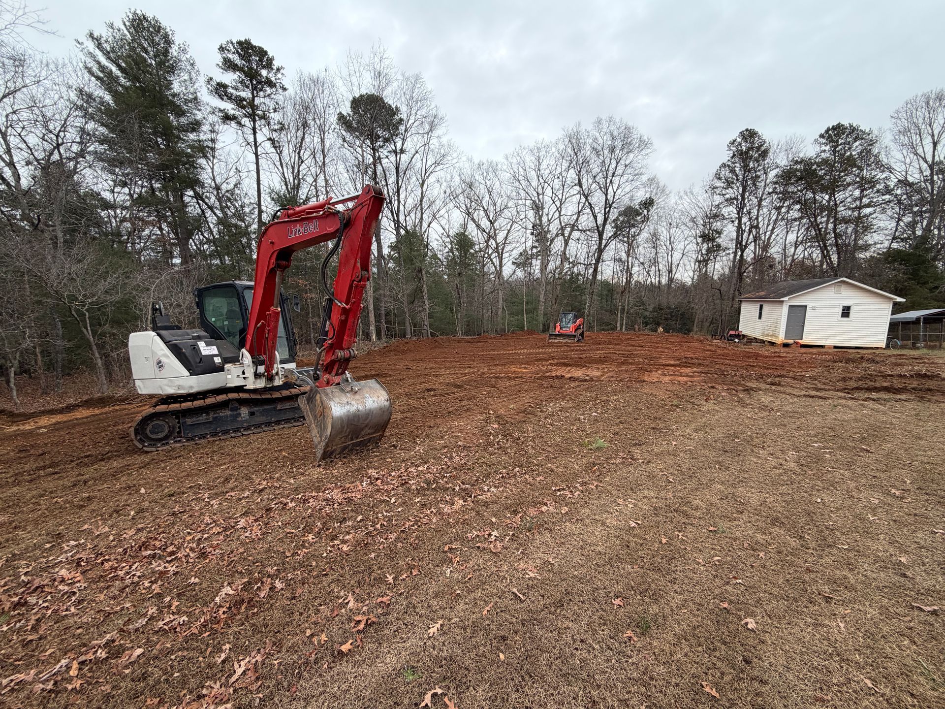 A red and white excavator parked on a cleared, muddy lot with a small white shed in the background.