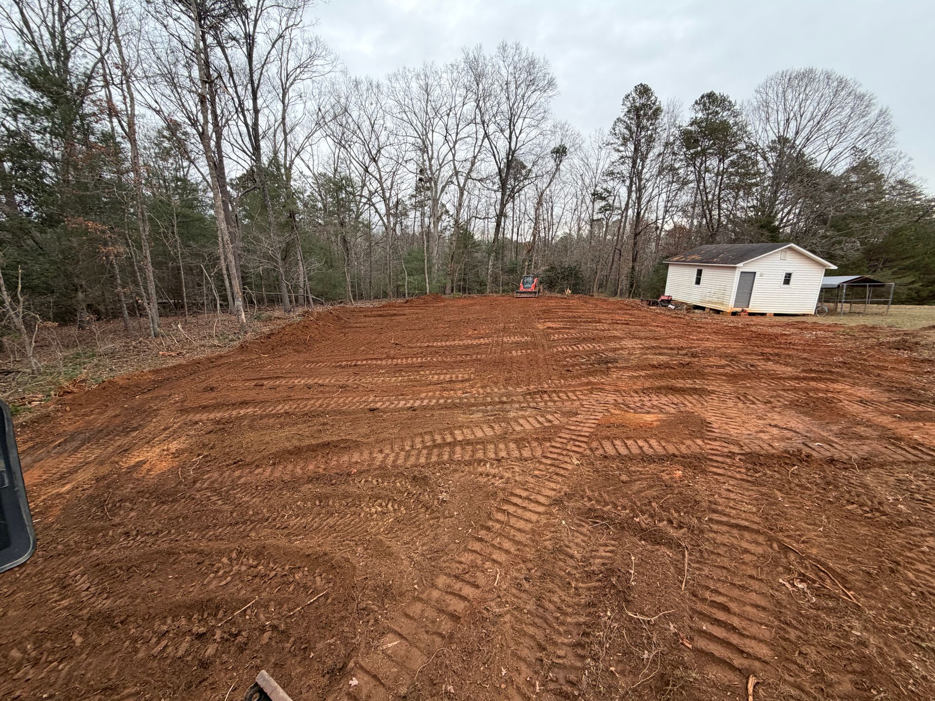 A large plot of freshly graded red dirt with tire tracks, situated in front of a small white house and wooded trees.