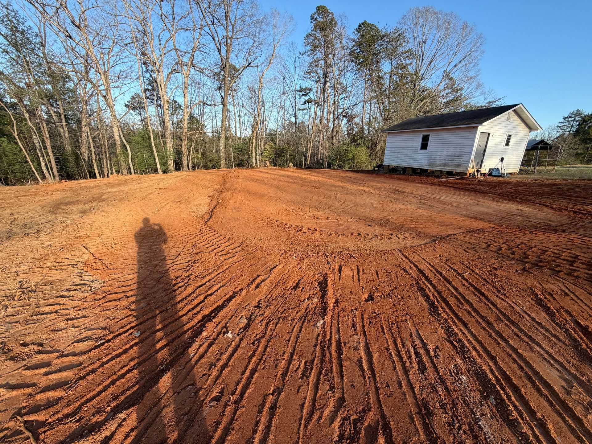 A wide, red dirt lot with heavy tire tracks in the foreground, next to a small, white, single-story house near trees.