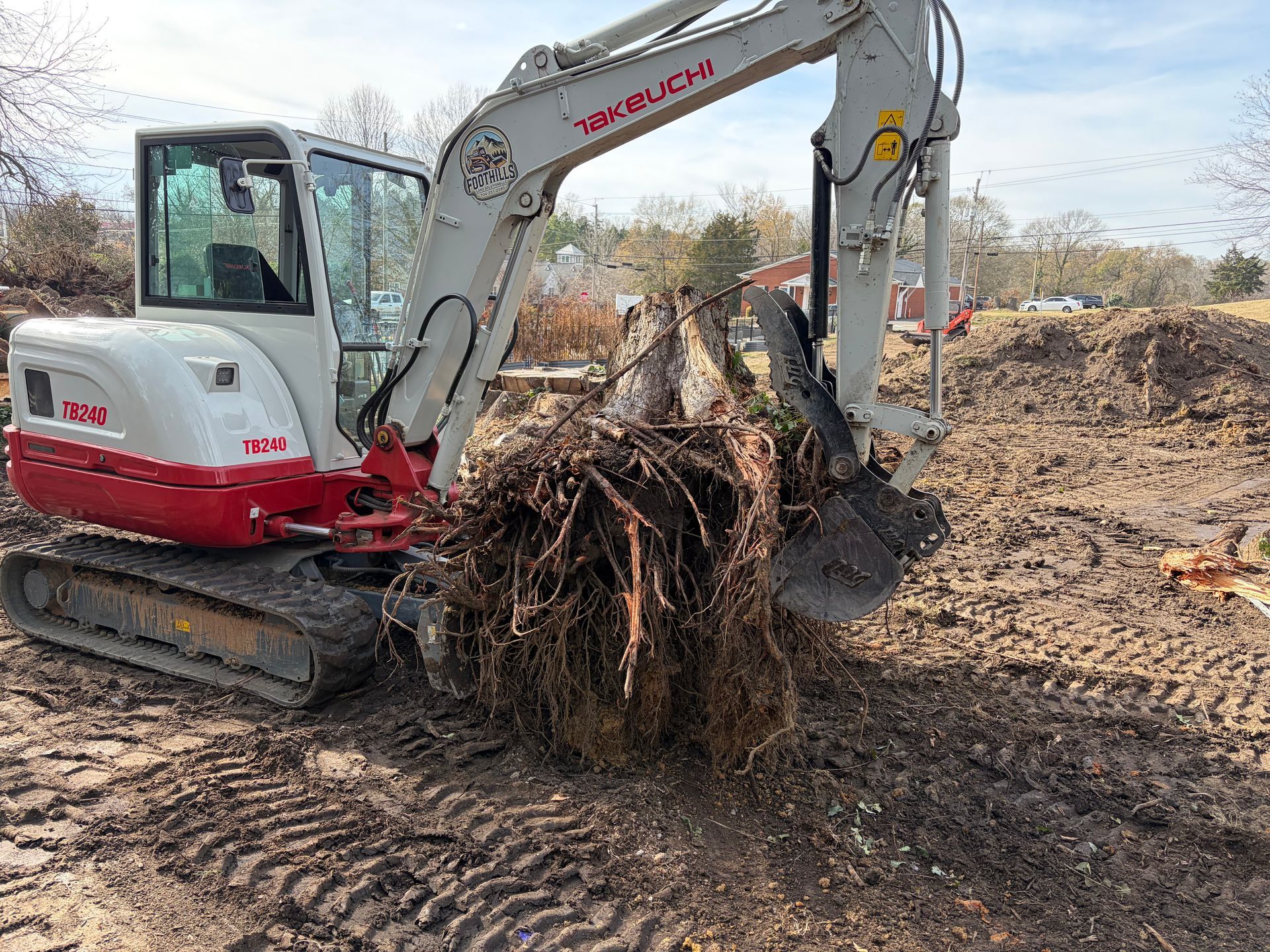 A white and red Takeuchi excavator sits on muddy ground, holding a large, uprooted tree stump with its bucket.