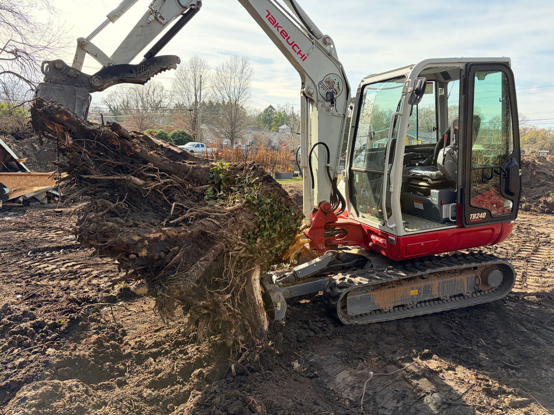 A white and red Takeuchi mini excavator lifting a large tree root ball from muddy ground on a sunny day.
