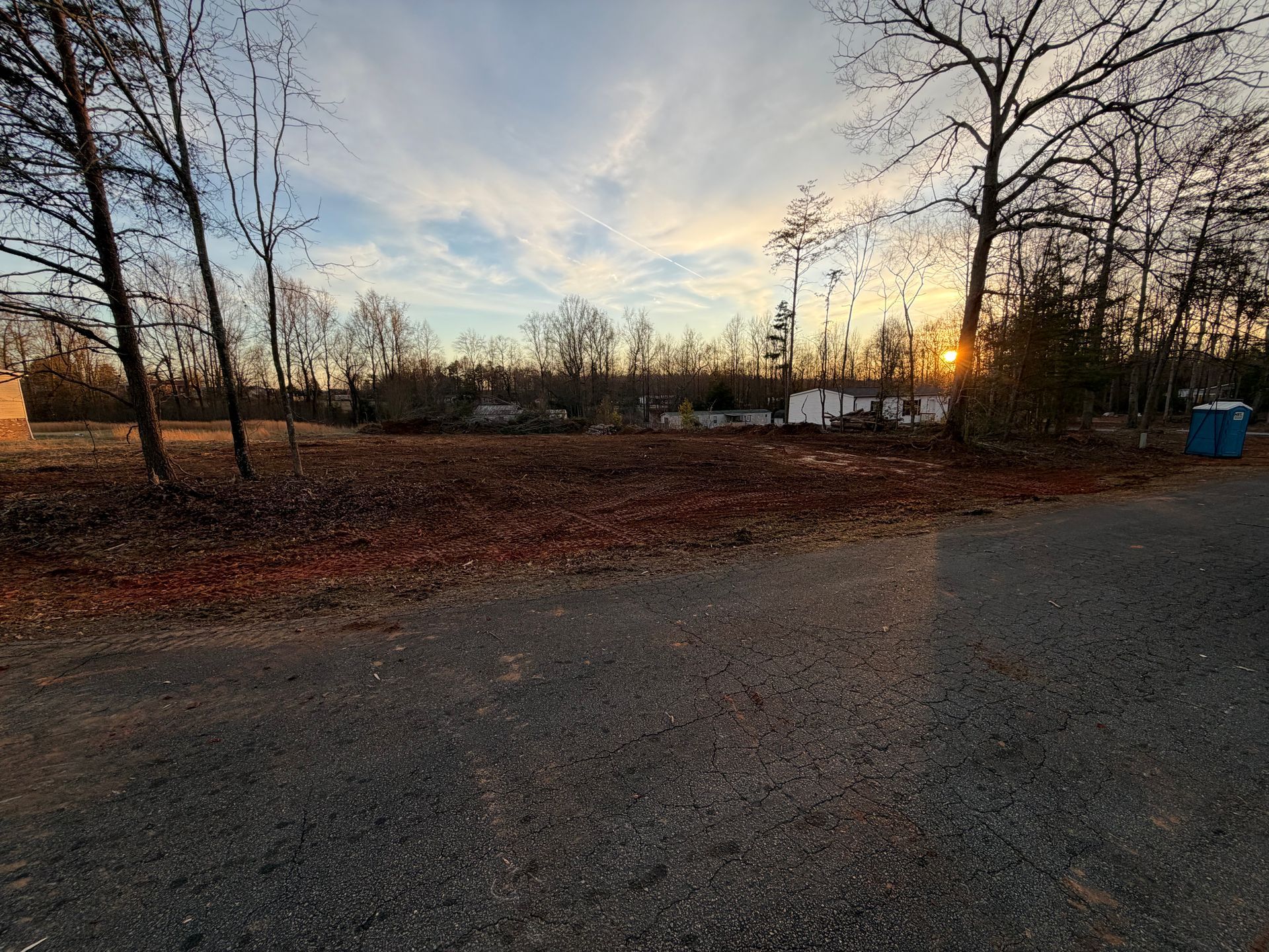 A cleared, leaf-covered lot at sunset with bare trees and distant buildings under a soft, light-filled sky.