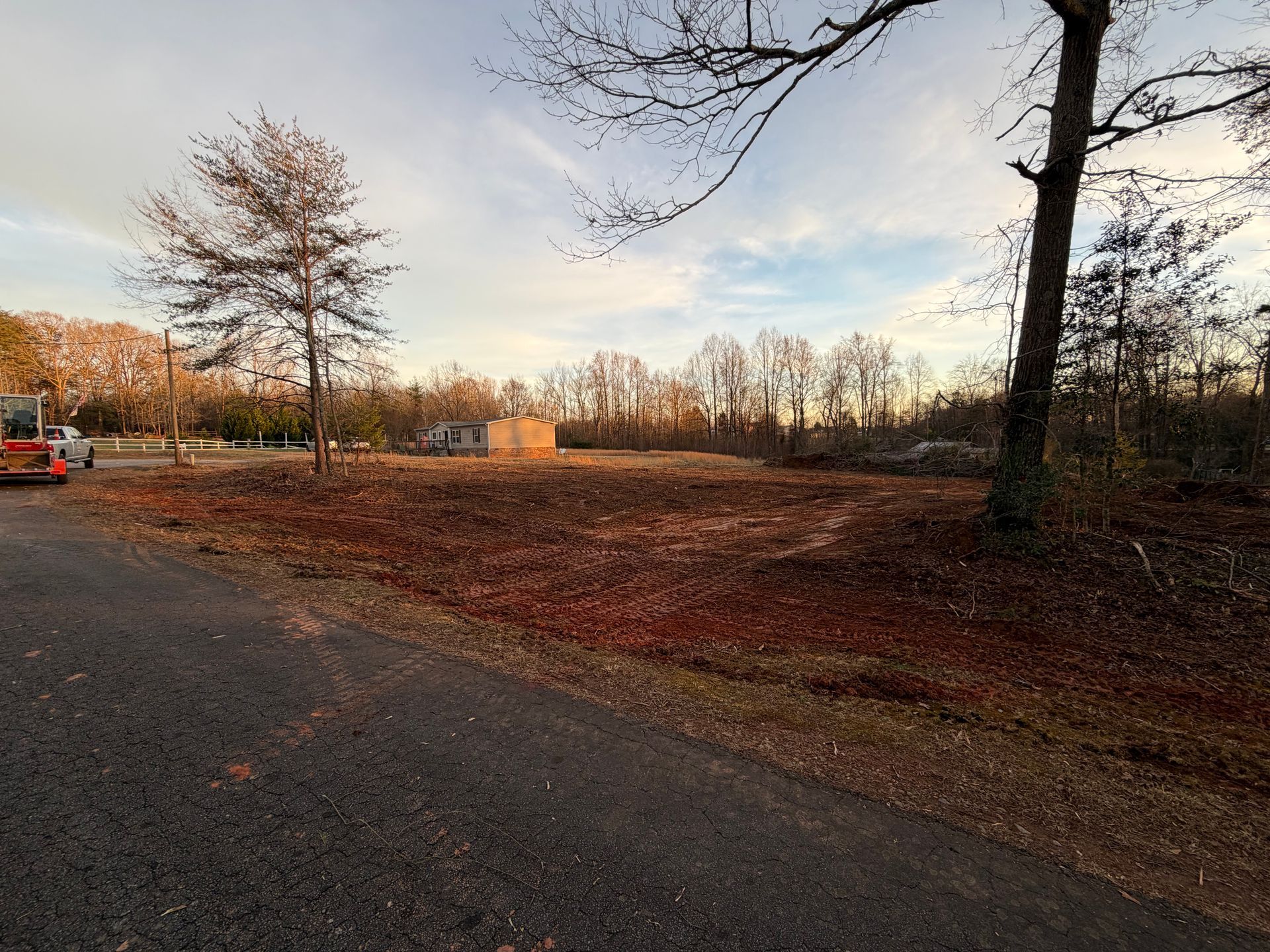 A clearing with scattered brown leaves in a wooded area, with a gravel road in the foreground and a distant building.