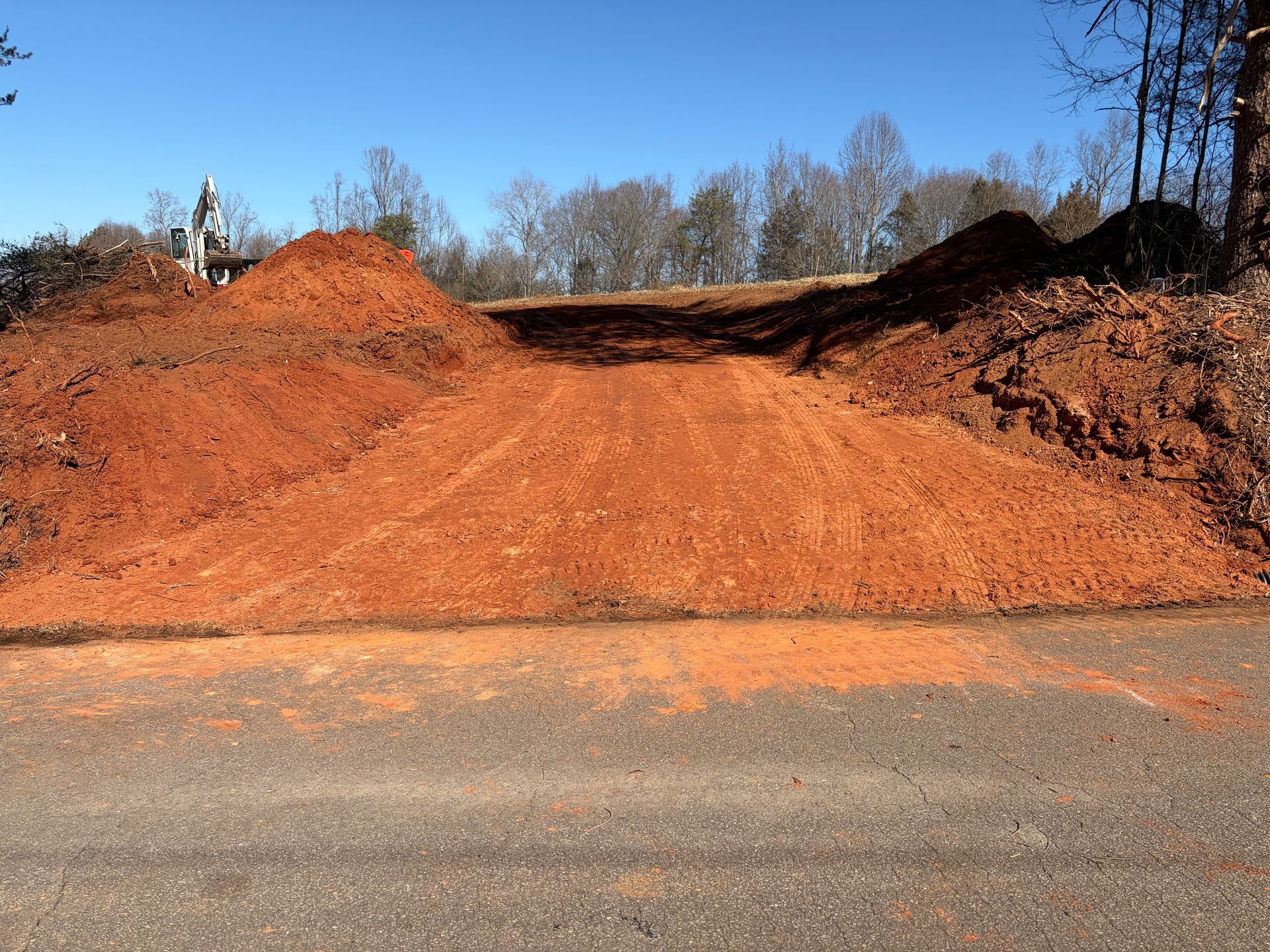 Red dirt mounds and a freshly cleared access road slope up from an asphalt road toward a wooded area under a blue sky.