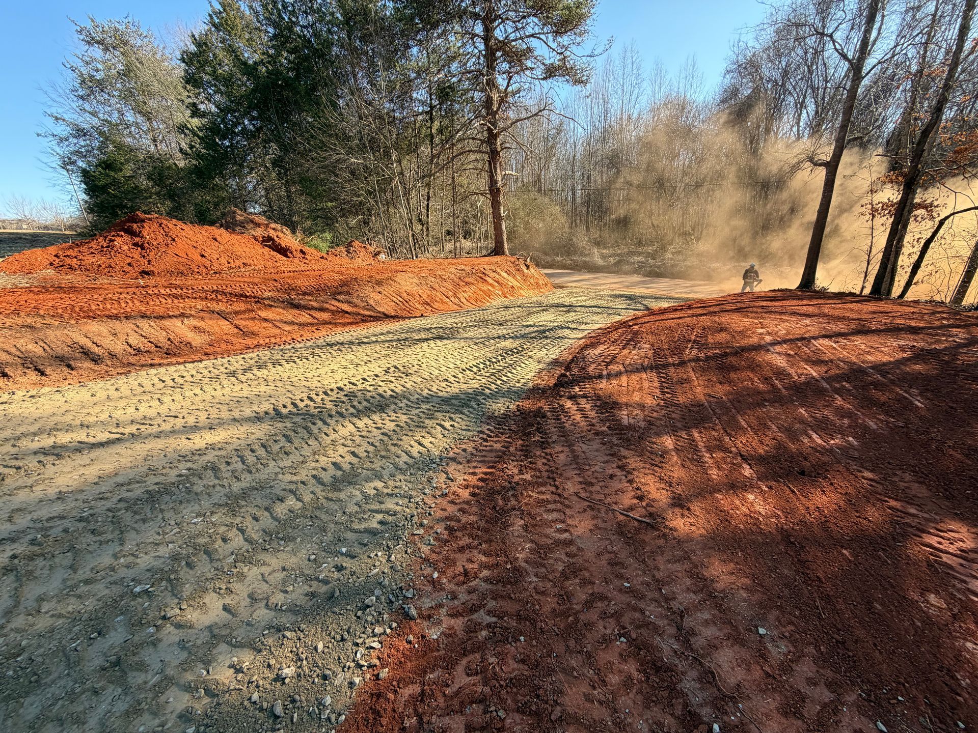 A gravel path winds through a construction site with piles of red dirt and trees under a clear blue sky.