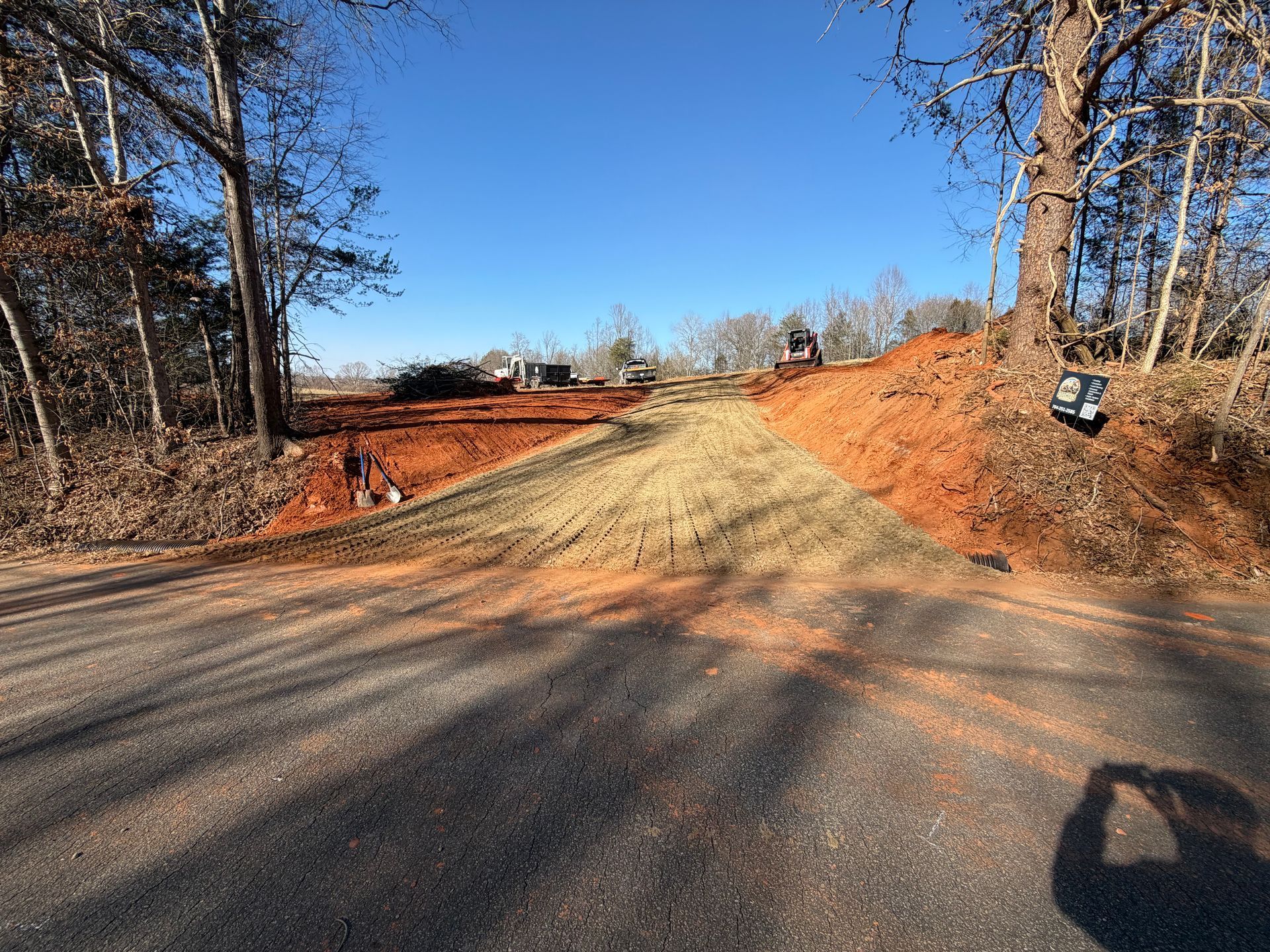 A gravel driveway access point being cleared at the edge of a paved road with red dirt banks and trees under blue skies.