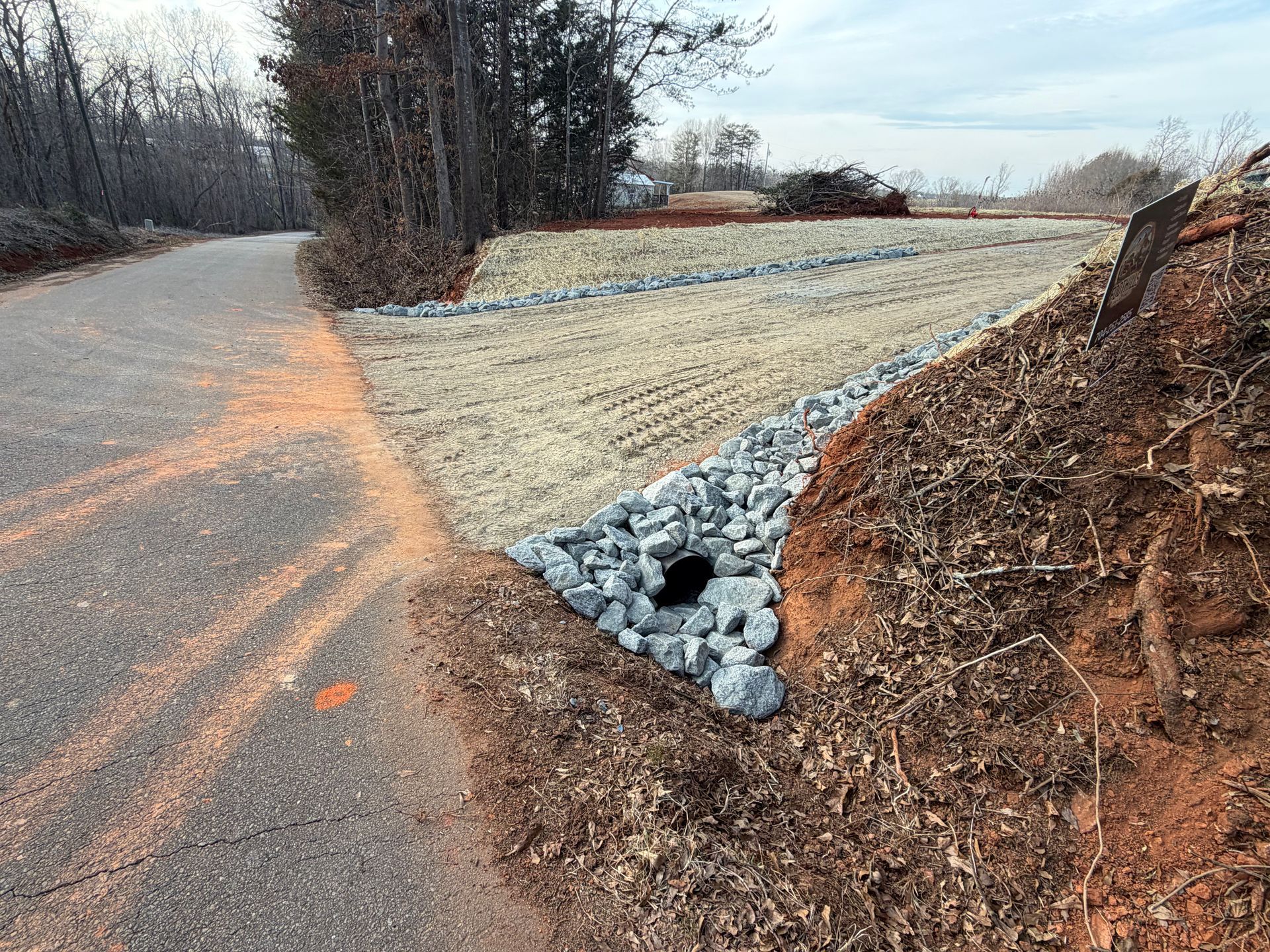 A paved road bordering a drainage ditch lined with grey rocks and light-colored straw mulch, with a culvert entrance.