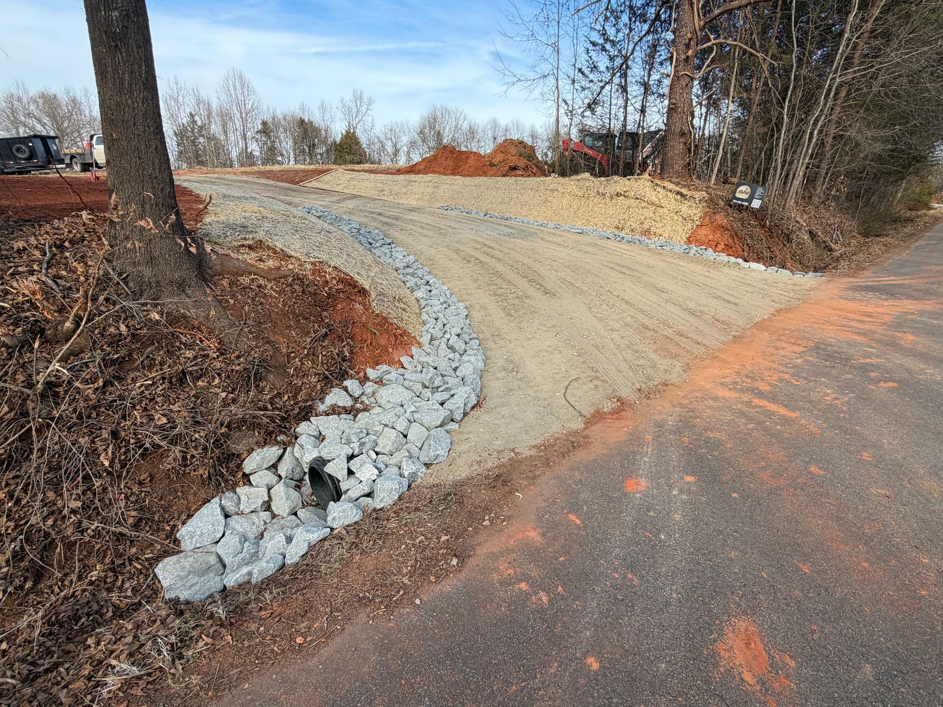 A dirt driveway entrance lined with grey riprap stone, next to a paved road and a dirt embankment with trees.