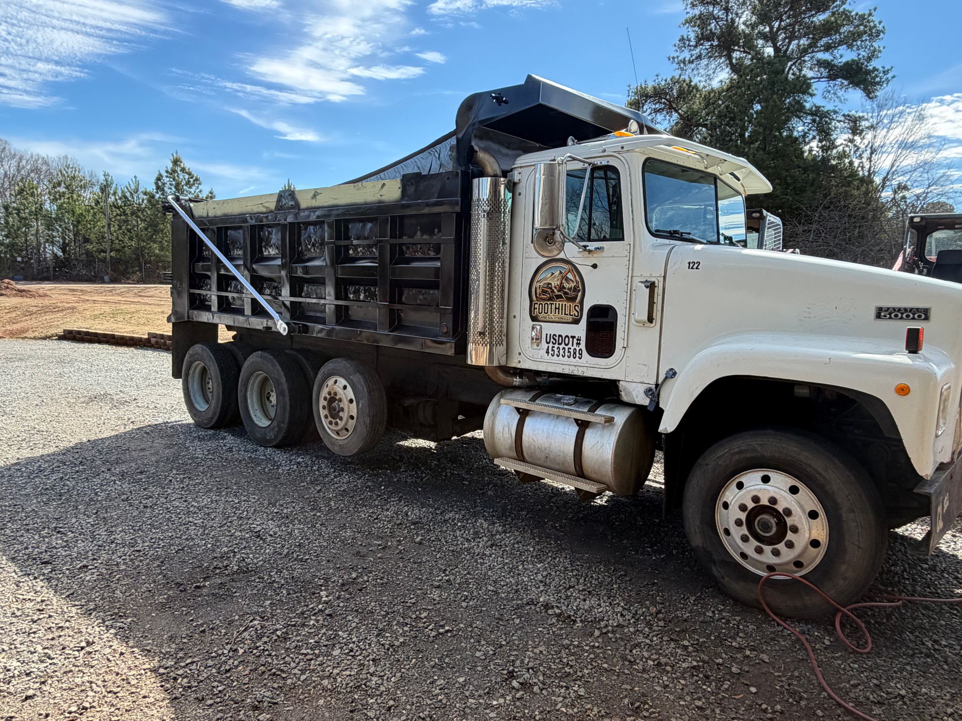 A white dump truck with a black cargo bed parked on a gravel lot under a bright, sunny sky.