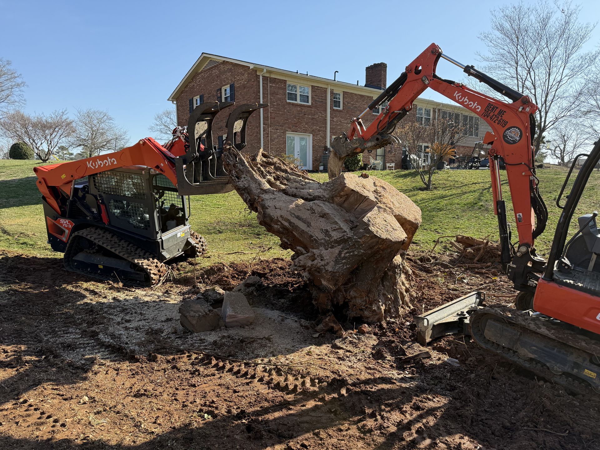 Two orange Kubota construction machines work together to lift and move a large tree stump in a yard near a brick house.