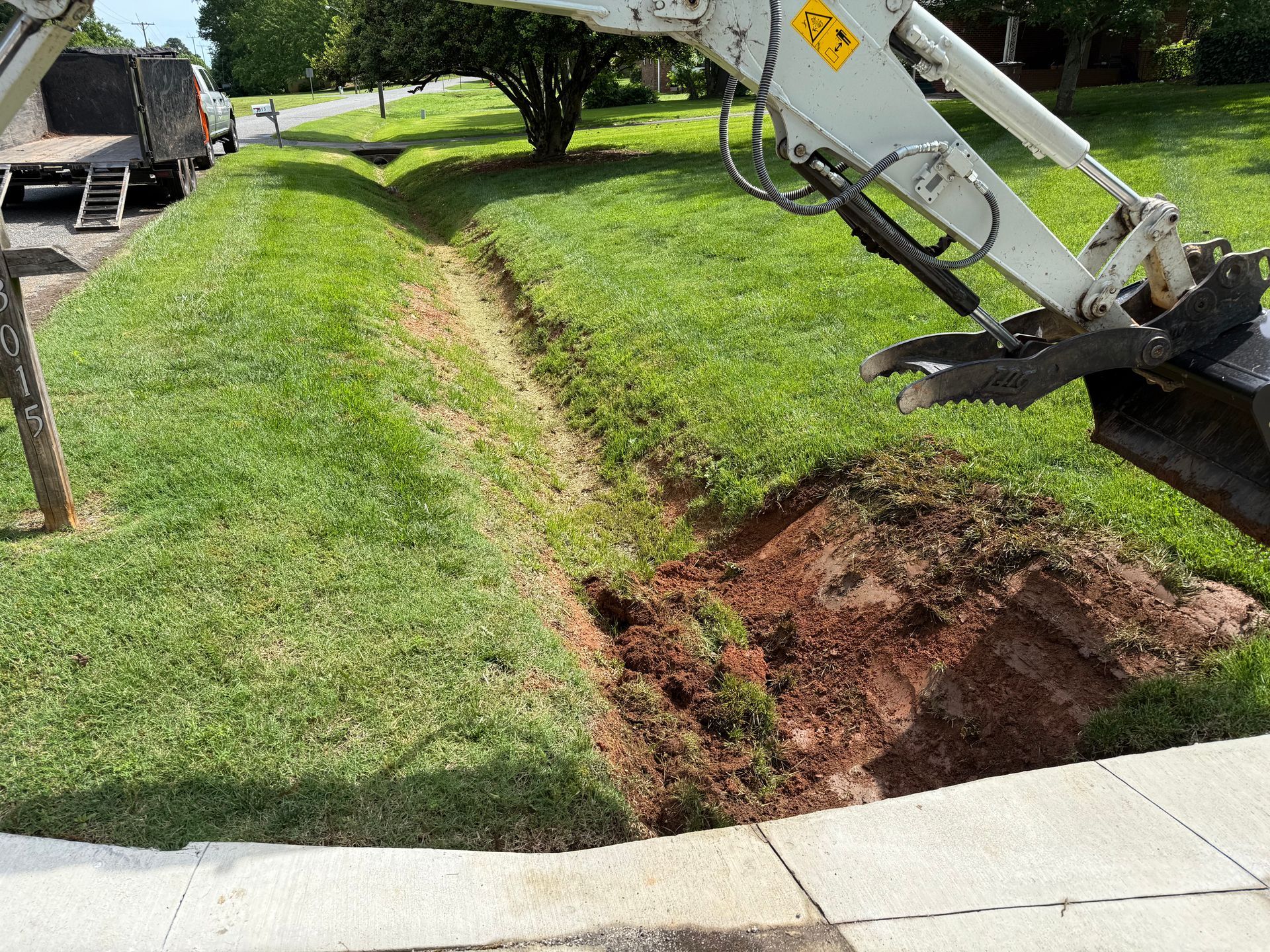 An excavator digging a trench in a residential lawn next to a sidewalk and a truck.