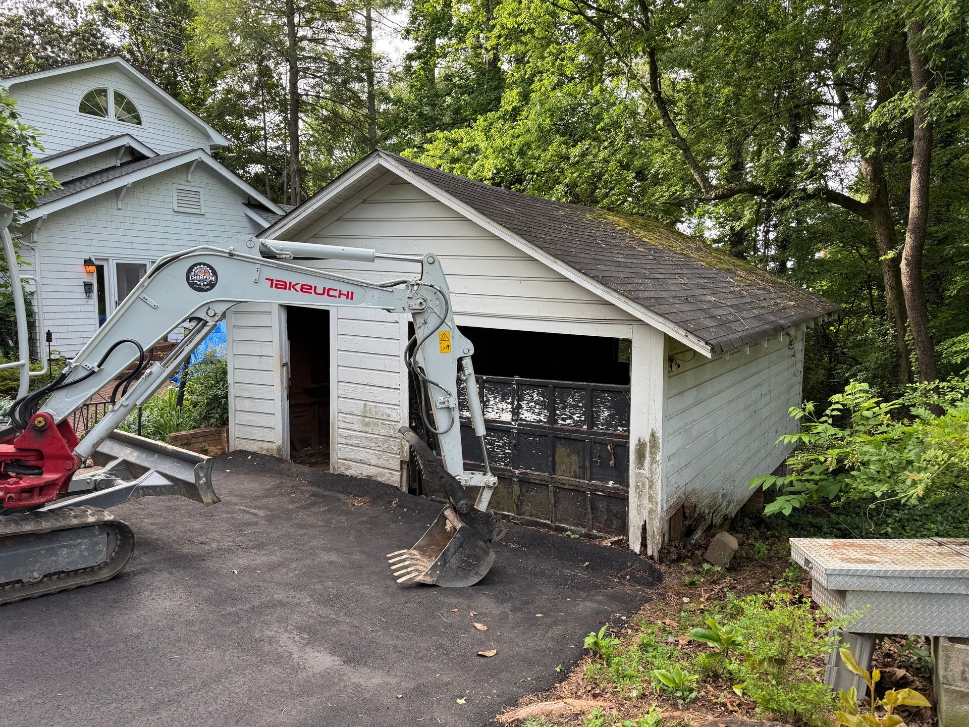 A white excavator parked in a driveway in front of a weathered white garage and a neighboring house.