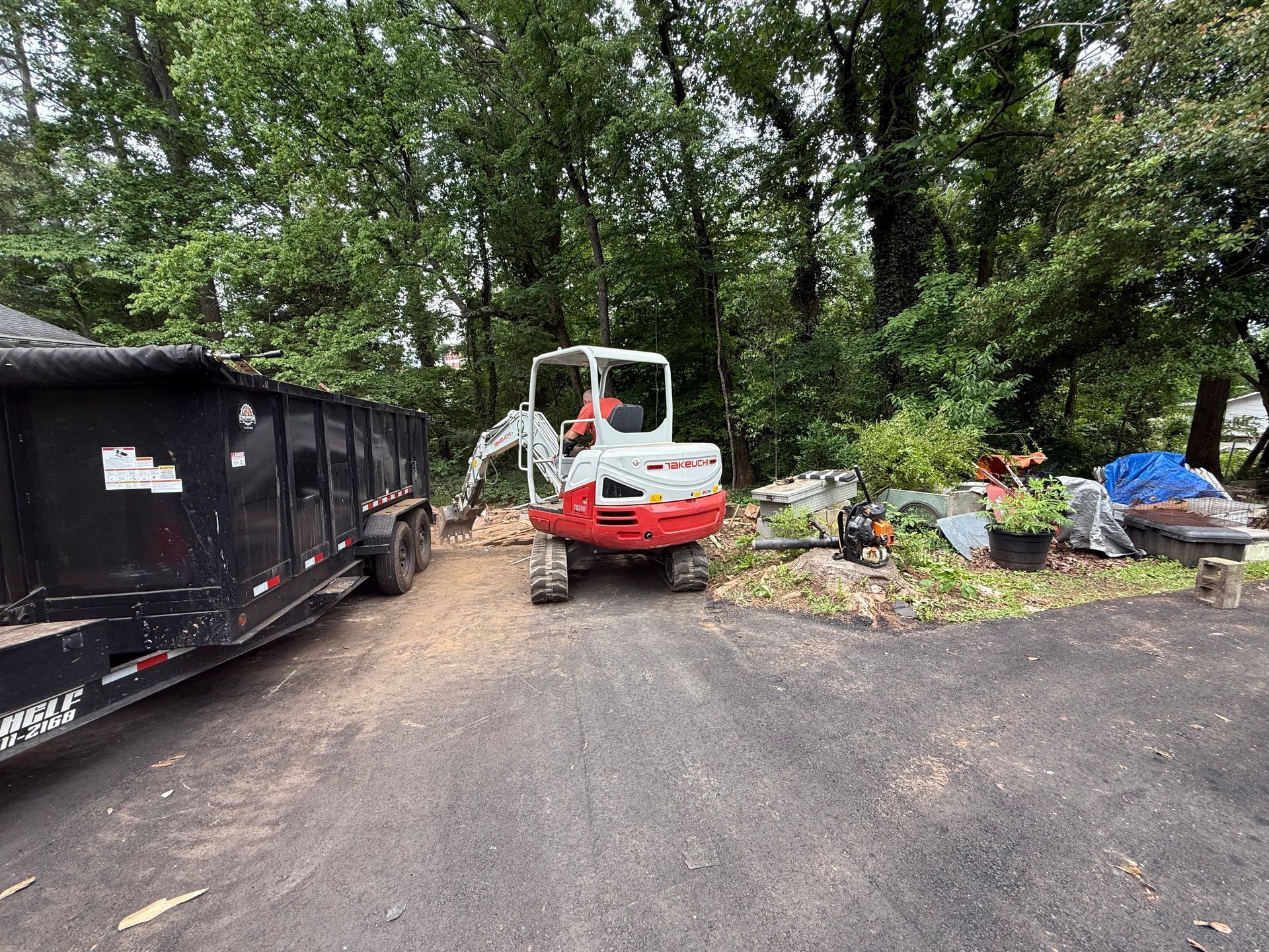 A white and red excavator parked next to a black dump trailer in a wooded residential driveway.