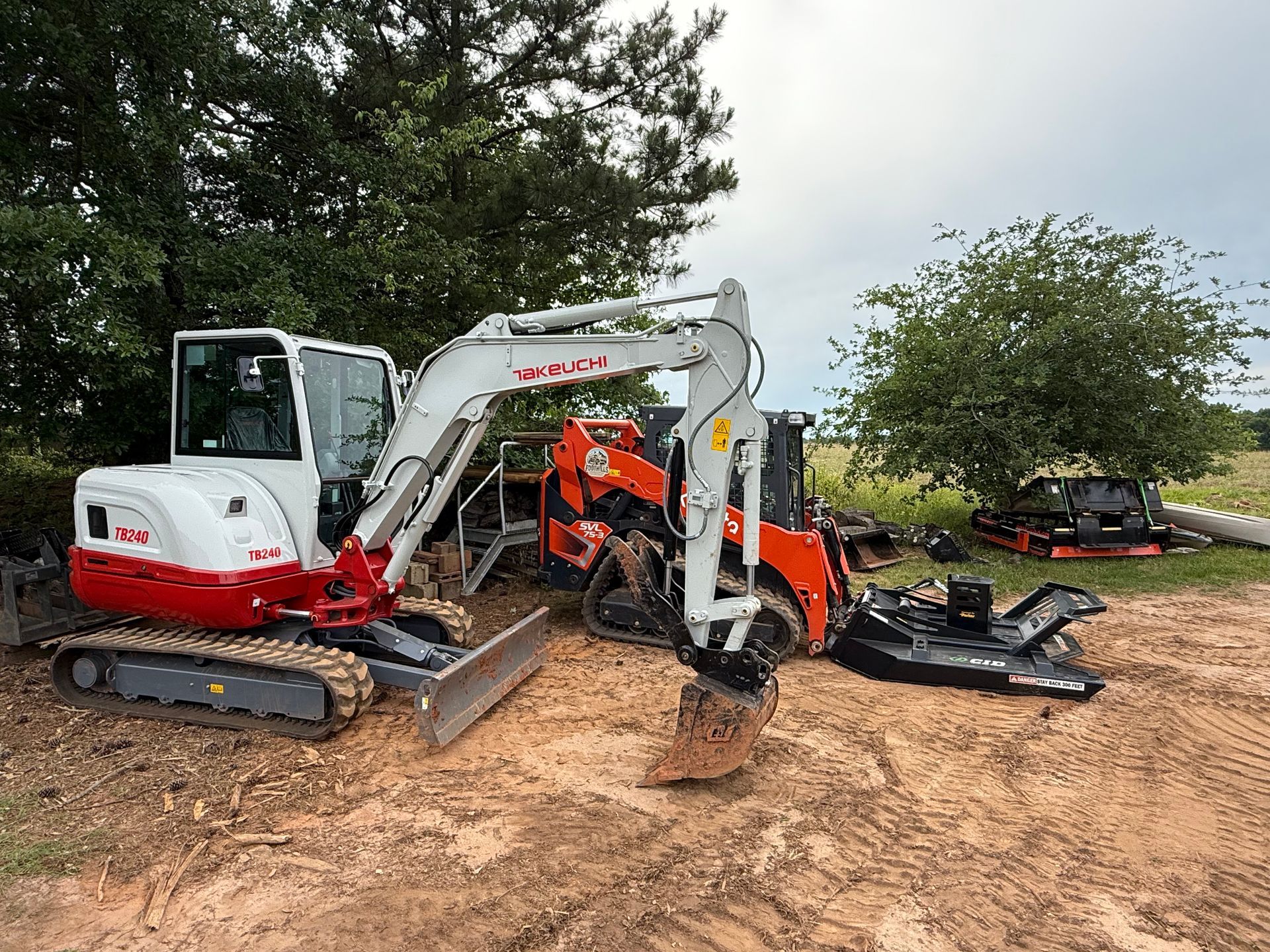 A white Takeuchi mini excavator sits on a dirt field with an orange skid steer and brush mower attachments nearby.