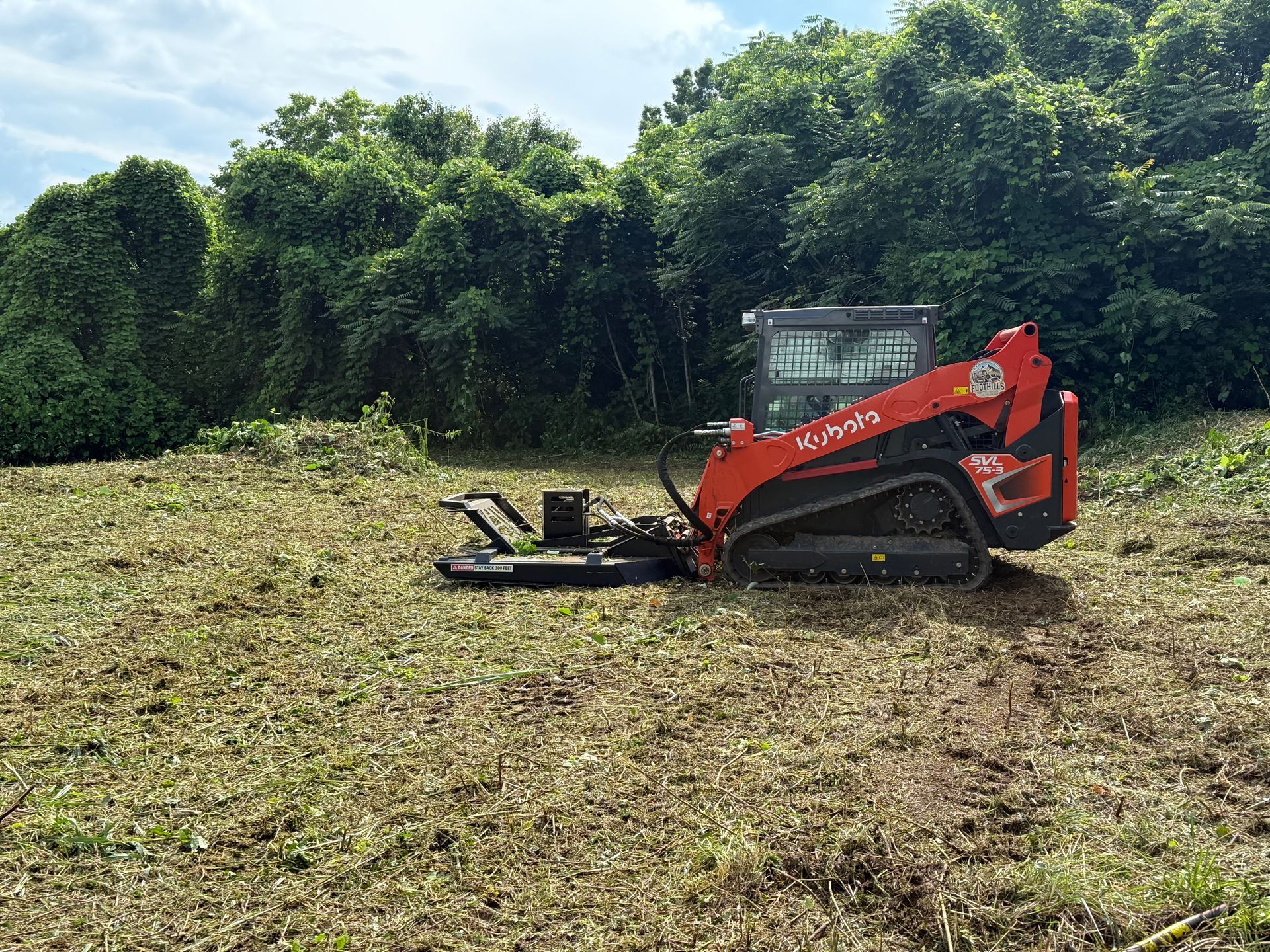 A red Kubota compact track loader with a brush cutter attachment works on a cleared field near a treeline.