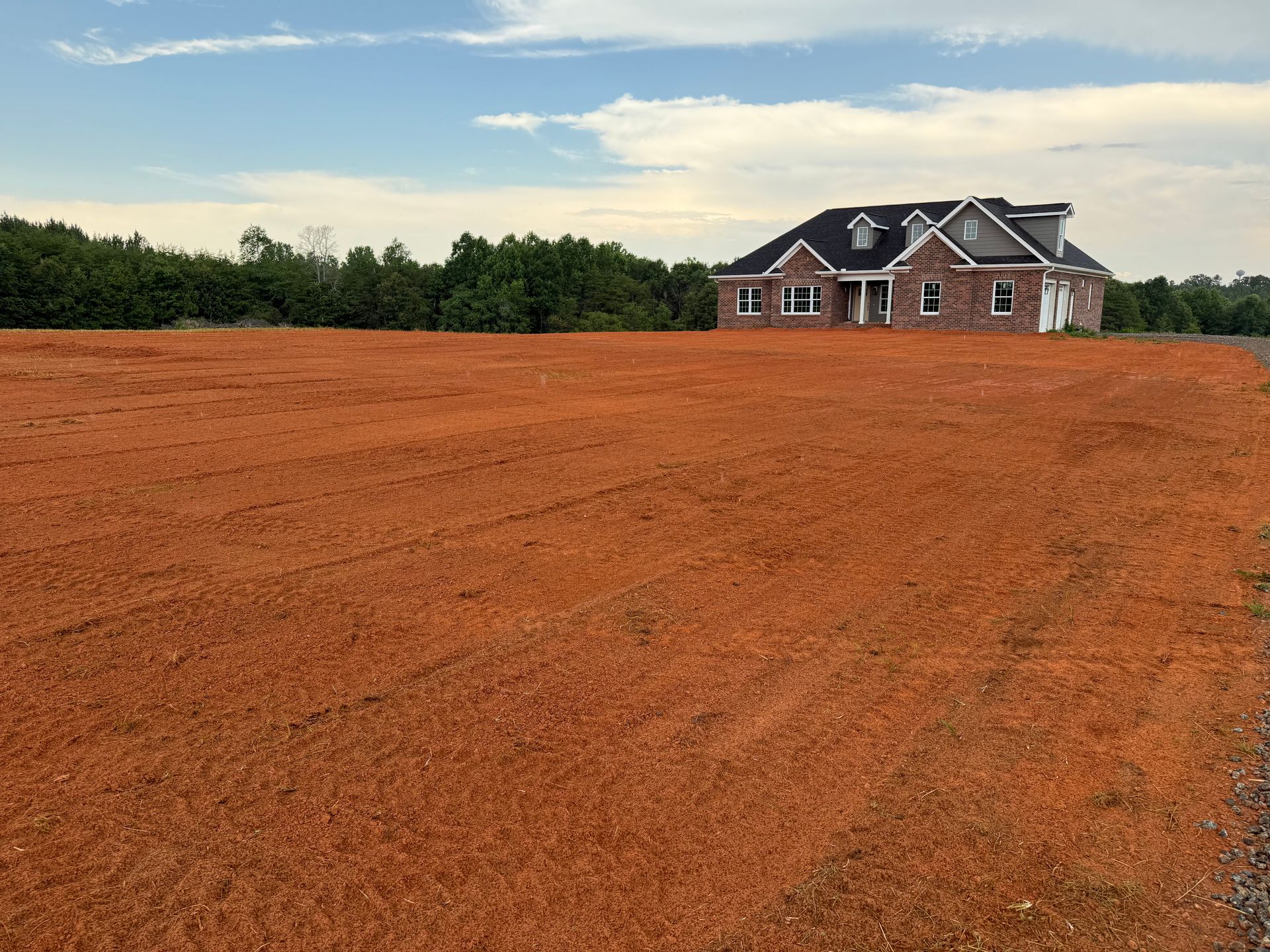 A house sits behind a large, leveled plot of reddish-brown dirt under a blue sky with soft white clouds.