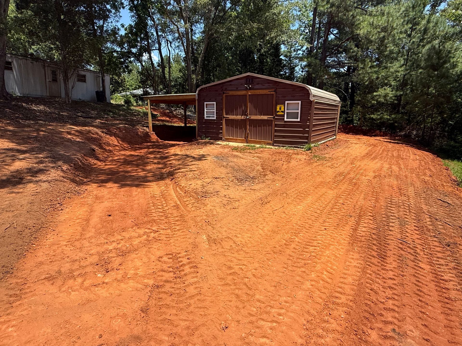 A brown storage shed sits on a cleared, red dirt lot surrounded by trees, with a small white building visible on the left.