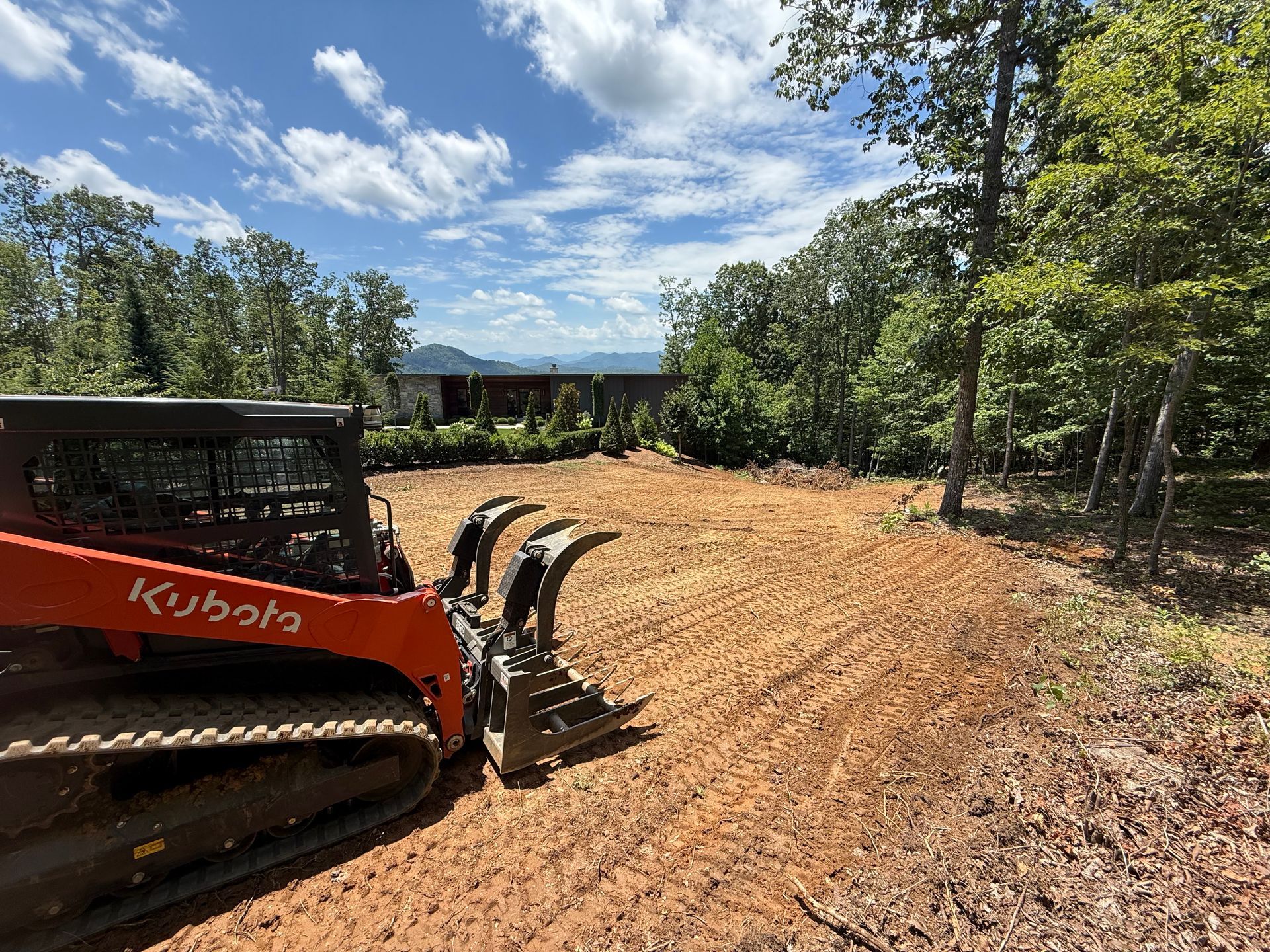 Orange Kubota compact track loader with a grapple attachment parked on a cleared, dirt lot near woods under a sunny sky.