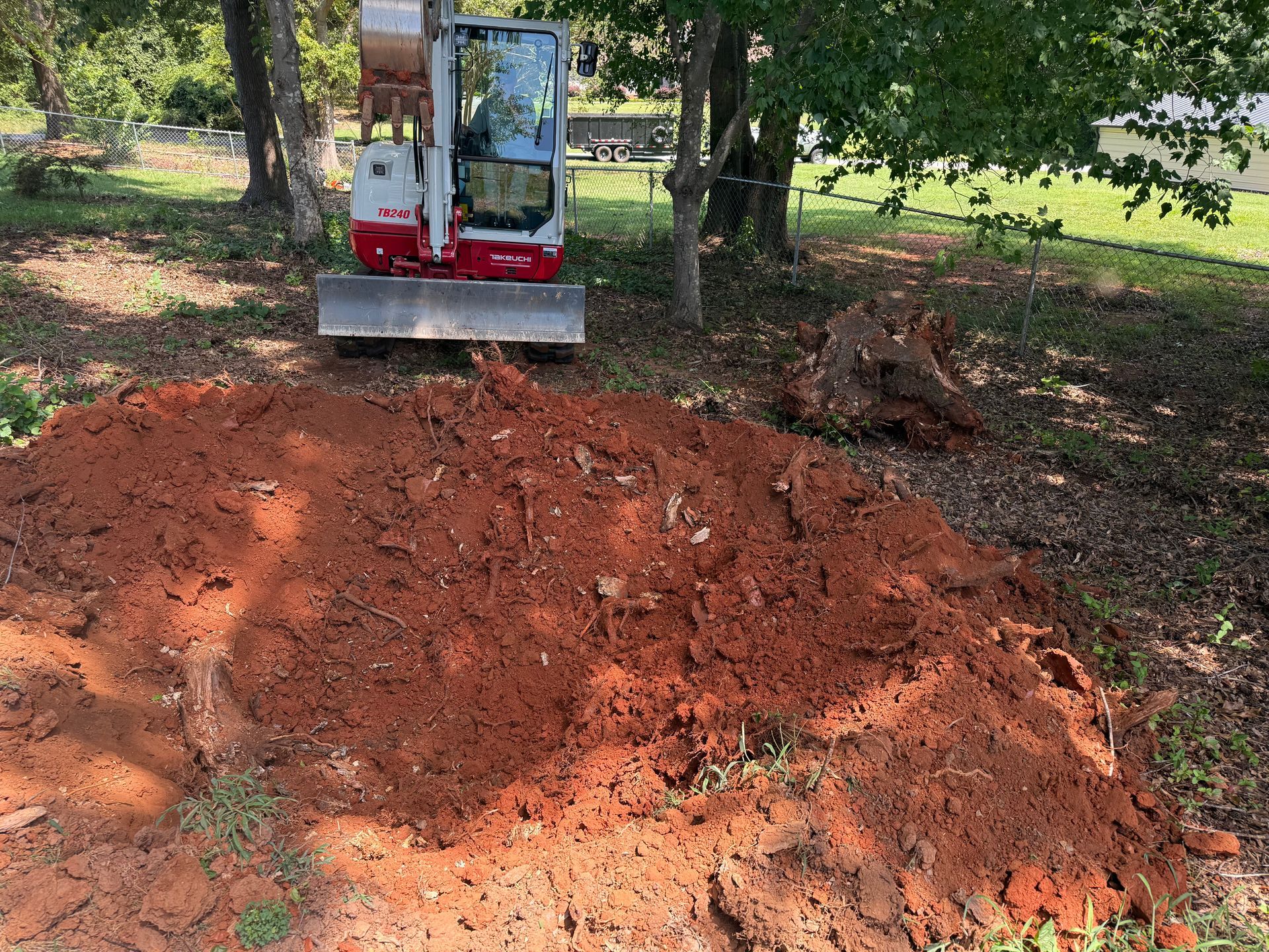 A small, white Takeuchi excavator sits parked behind a large, freshly dug pile of red clay in a grassy, shaded yard.