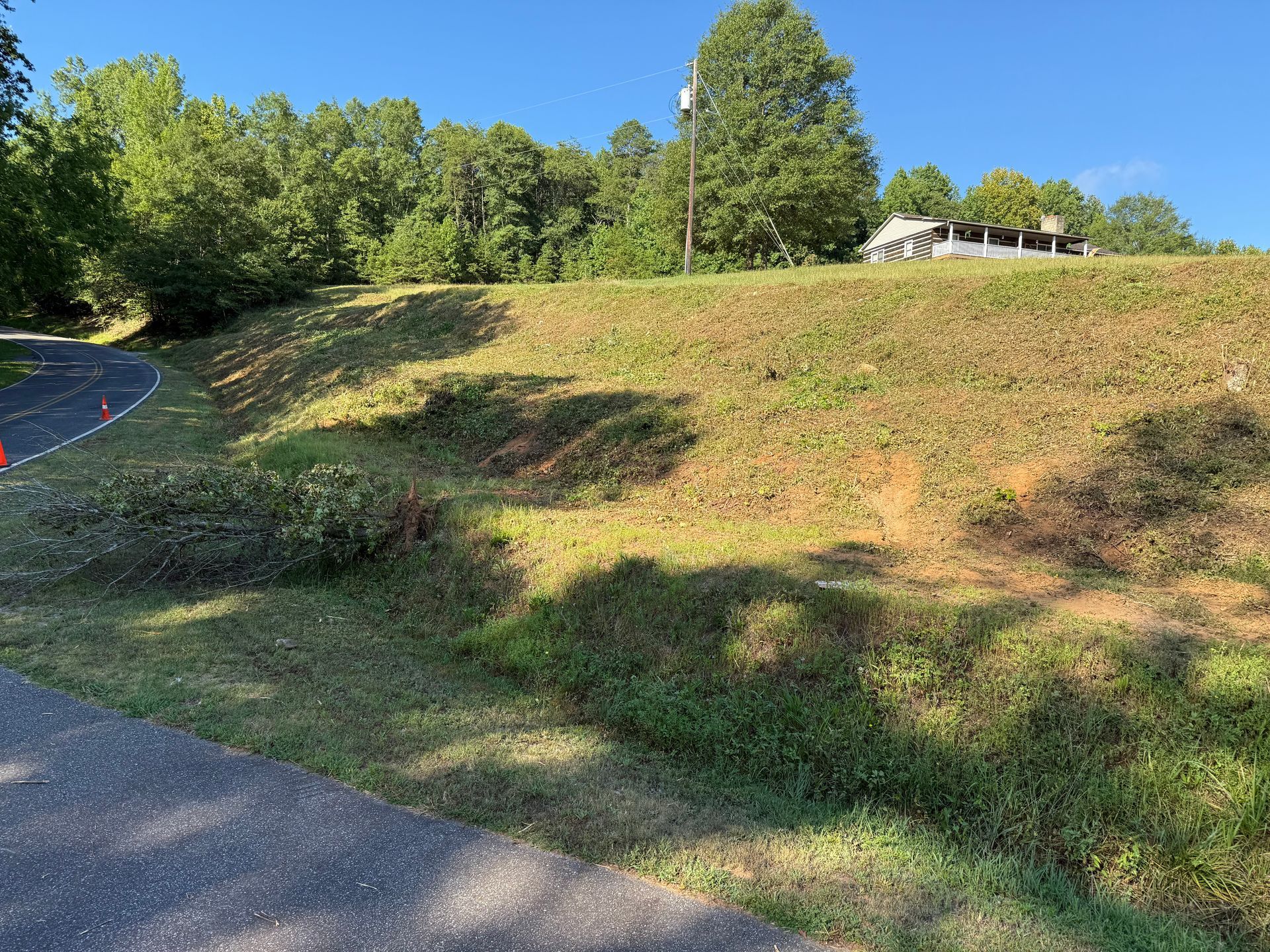 A paved road curves past a grassy, sloped hill with scattered debris, leading toward trees and a building in the distance.