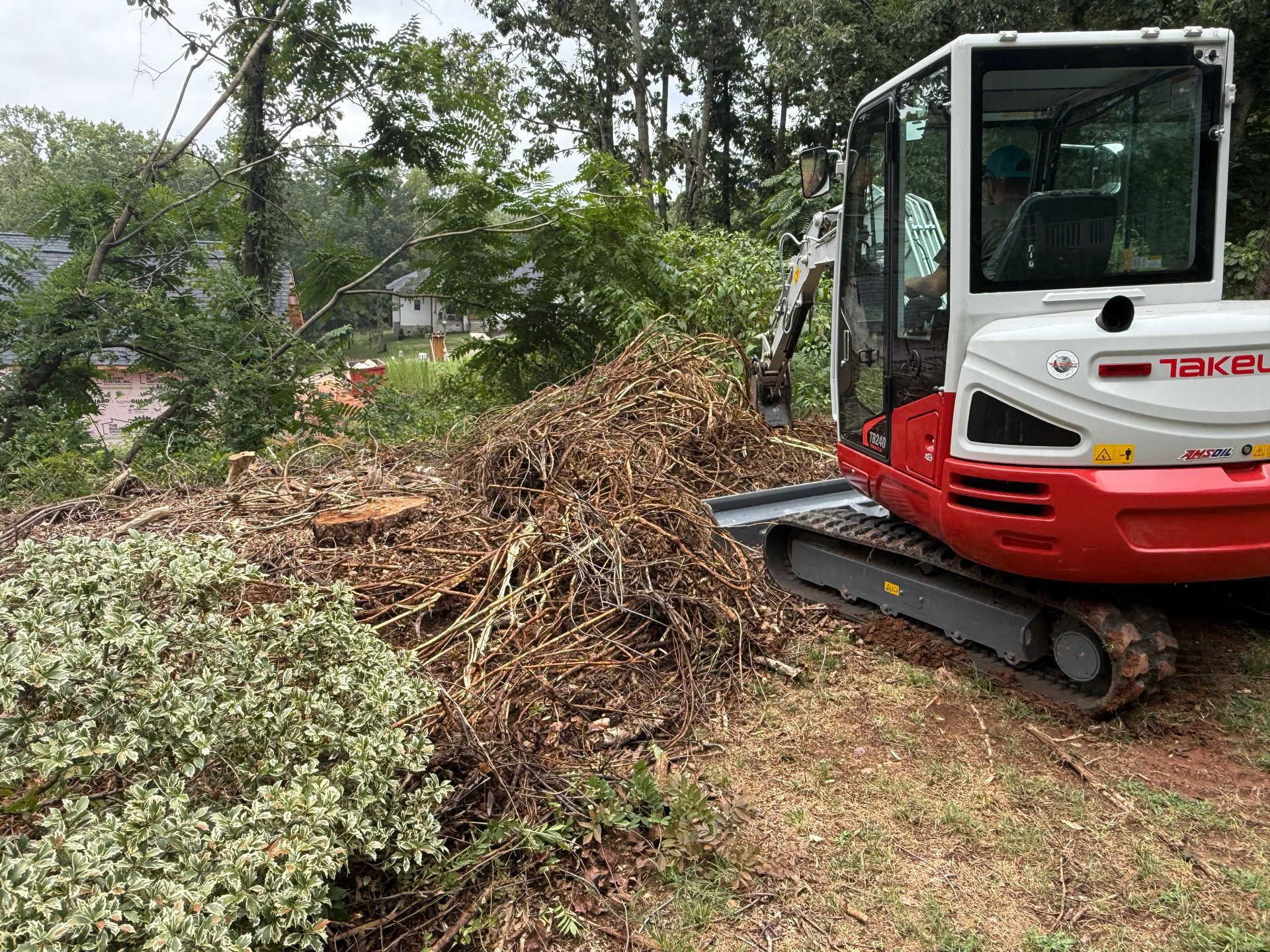 A red and white Takeuchi compact excavator sits next to a large pile of brush and debris in a wooded outdoor area.