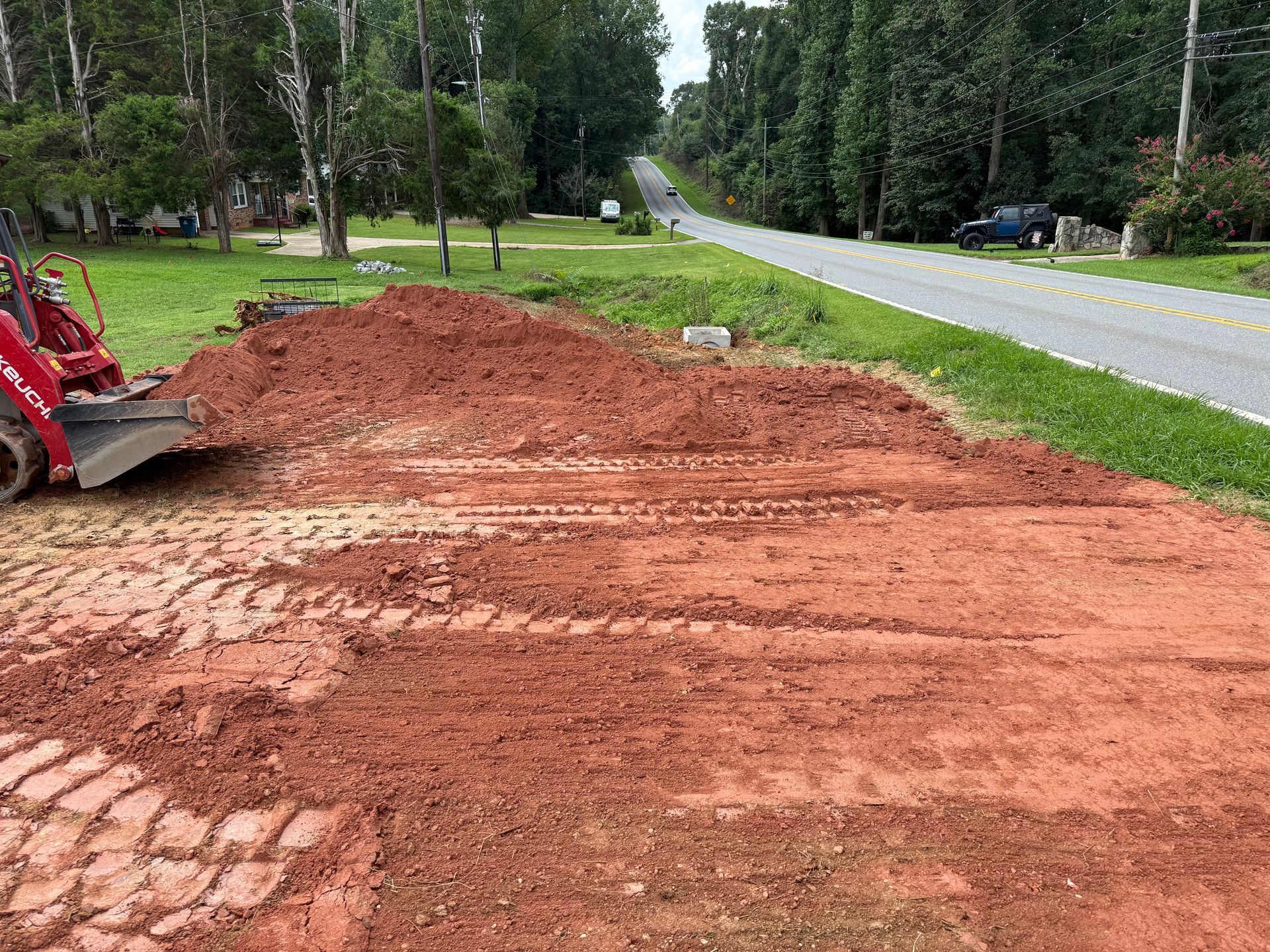 A red skid steer loader parked on a construction site with a pile of red dirt next to a road.