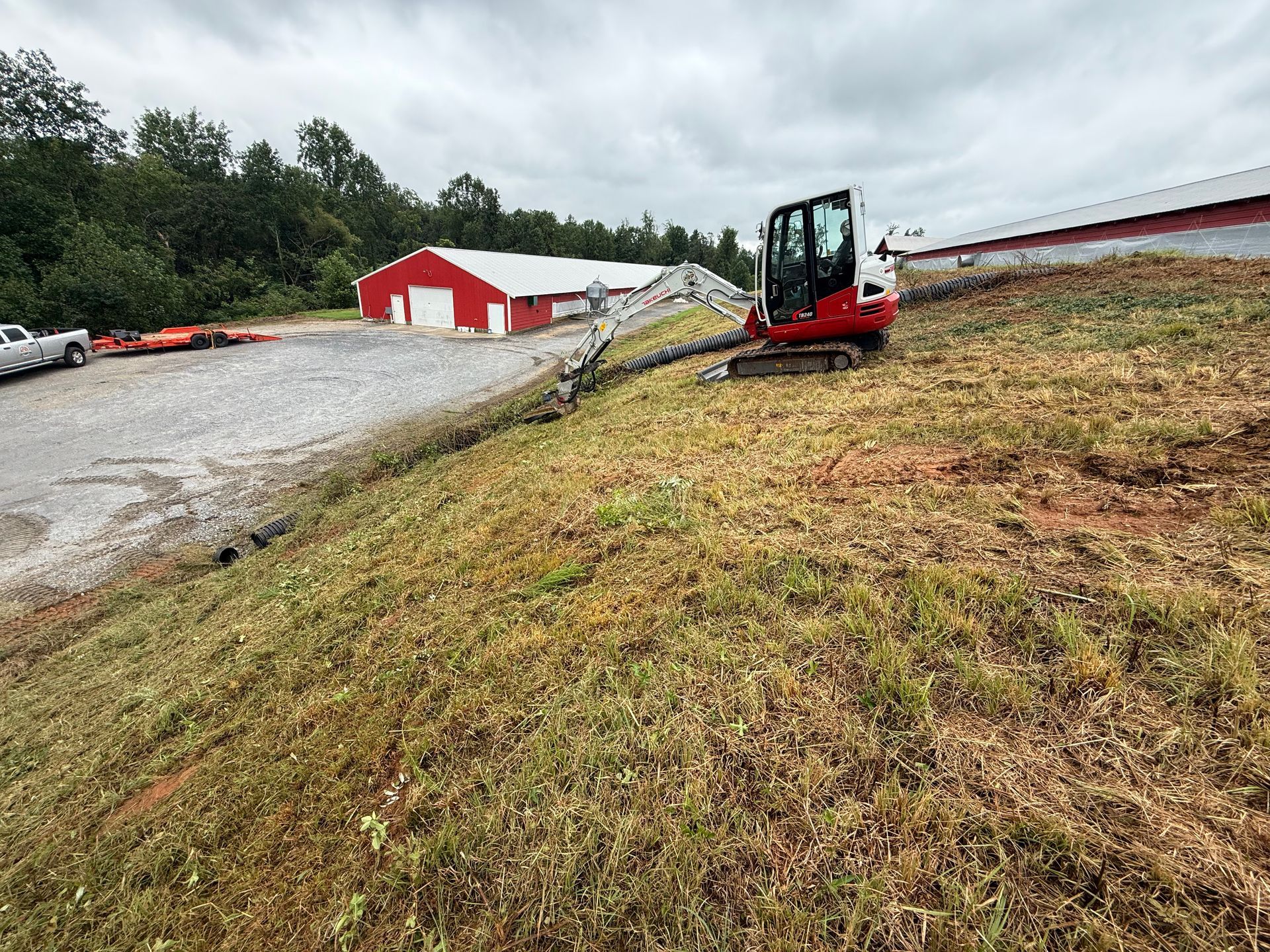A red mini excavator parked on a grassy hillside next to a gravel lot and farm buildings under a cloudy sky.