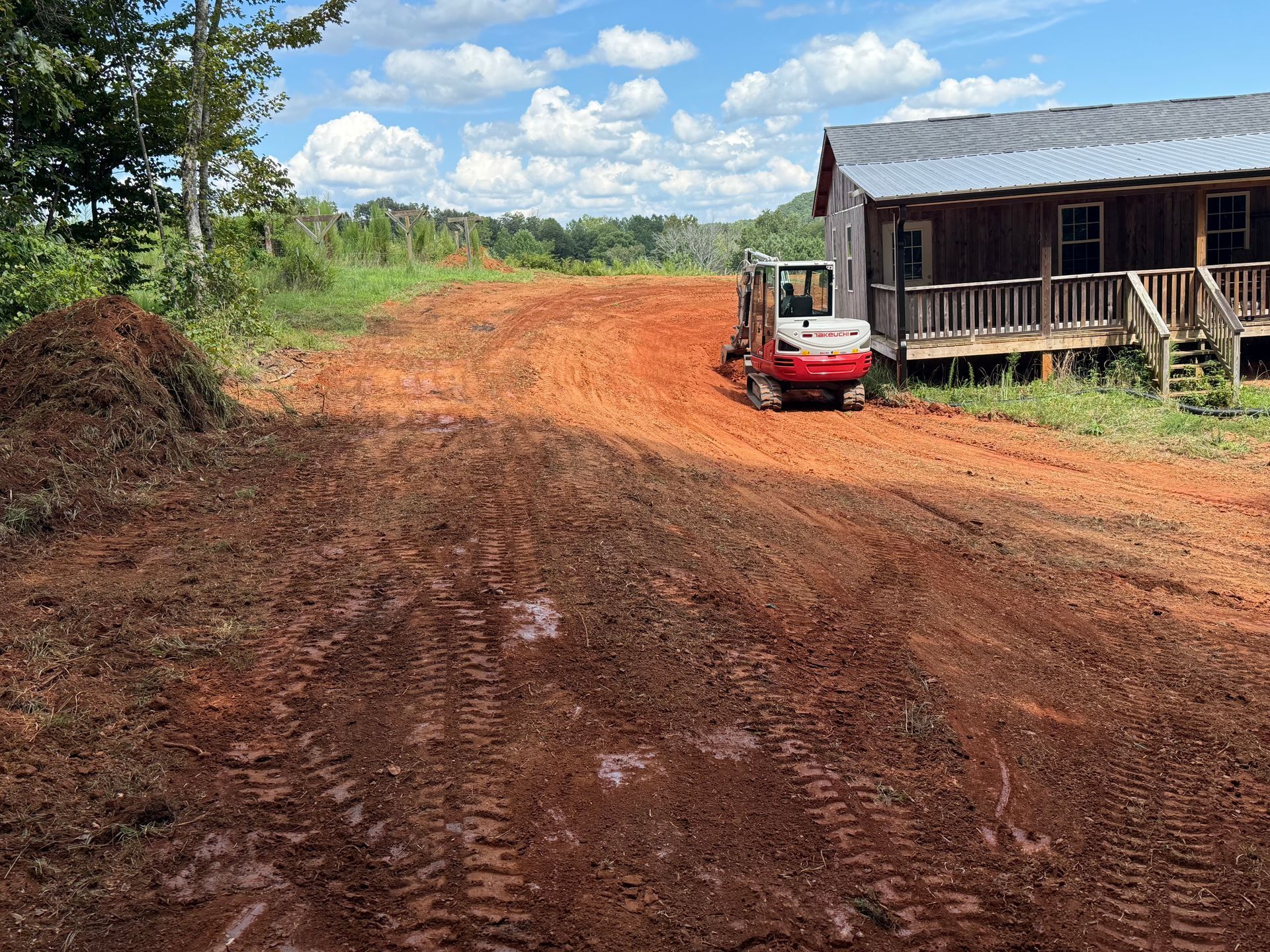 A small red construction excavator parked on red clay dirt near a rustic wooden house under a bright blue sky.