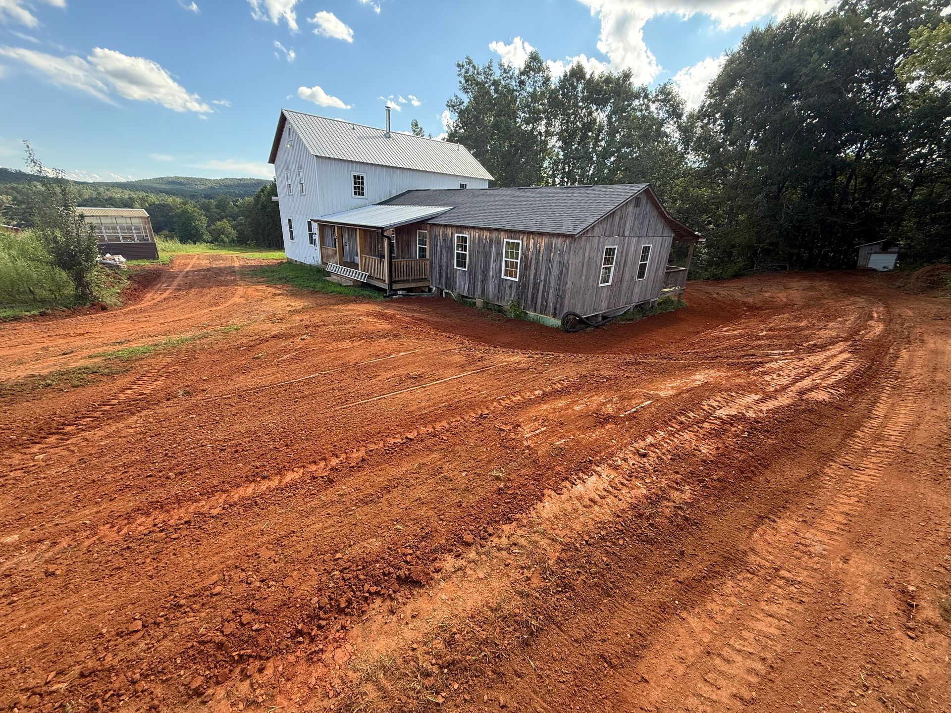 A white house attached to a weathered wooden structure, set amidst a large, freshly tilled red clay field.
