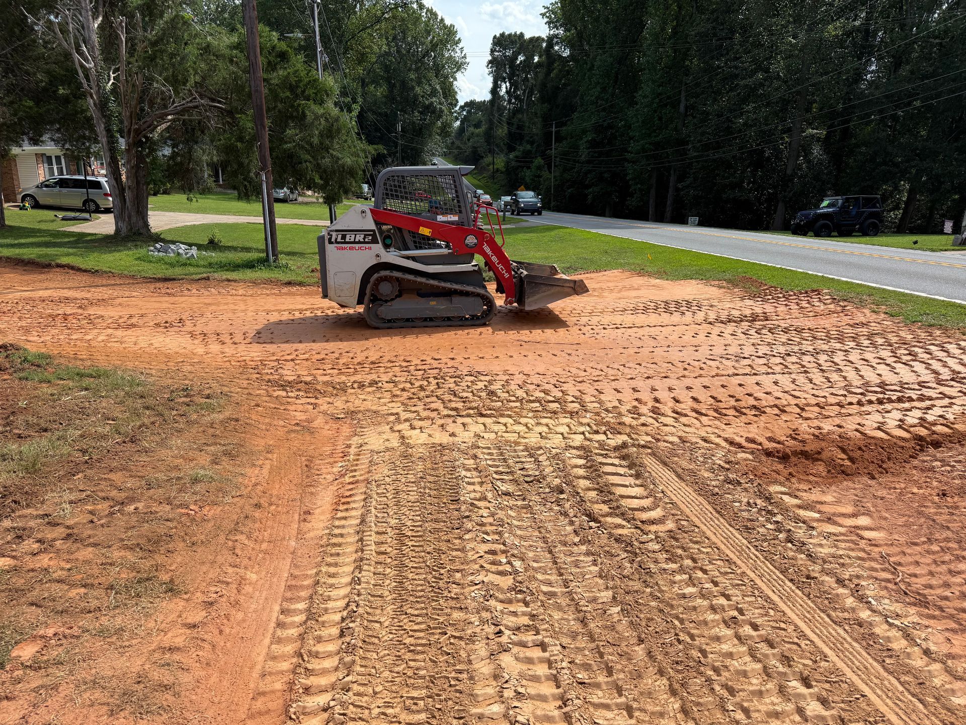 A red and white compact track loader sits on a freshly graded red dirt construction site beside a road and grassy area.