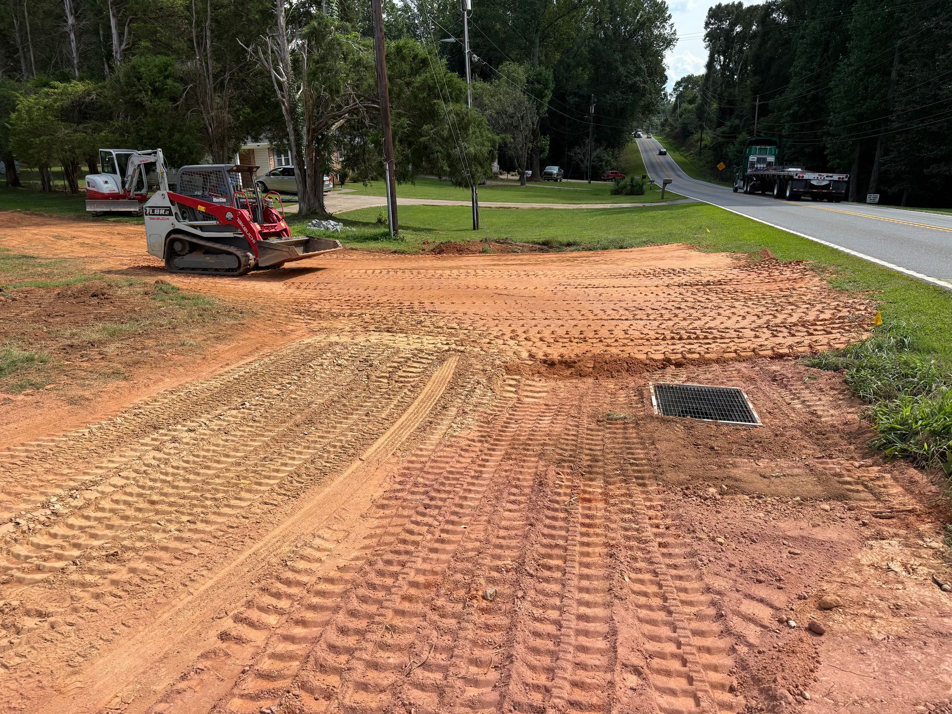 A red and white skid-steer loader sits on a cleared dirt lot next to a road, with a square storm drain visible.