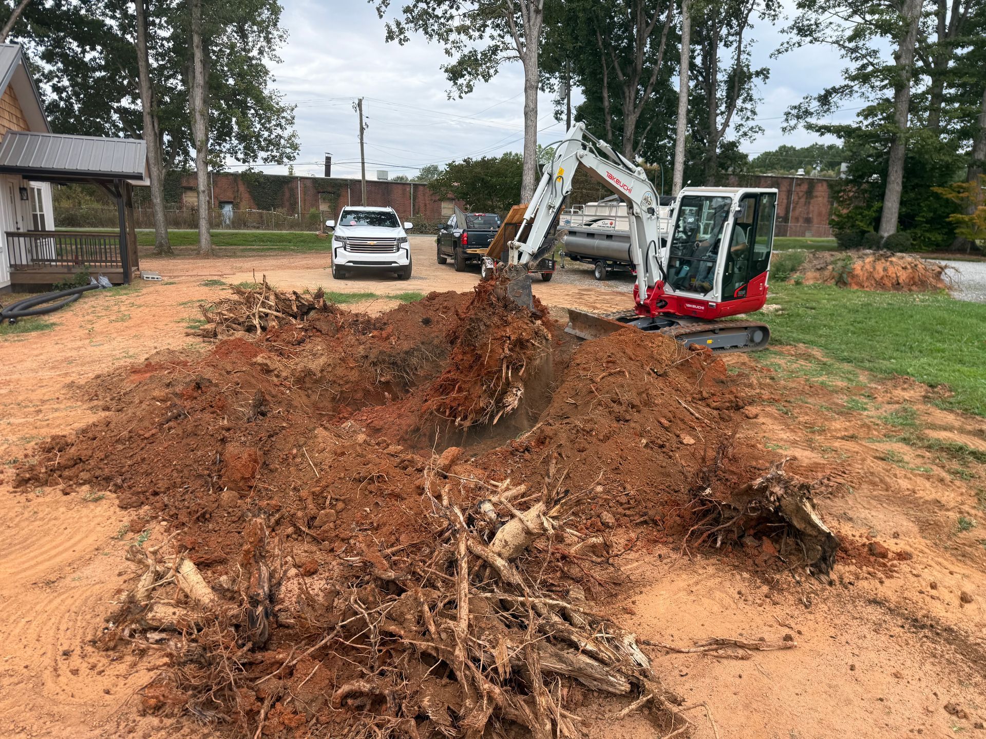 A red and white mini excavator digging through a large pile of dirt and tree roots in a grassy yard.