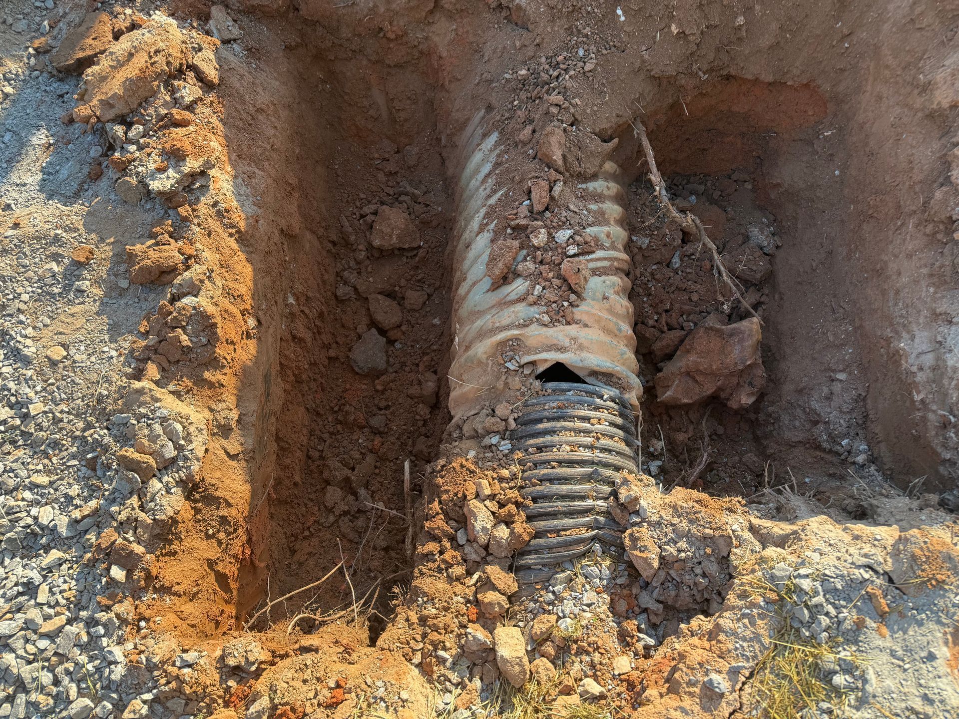 A dirt trench in the ground exposes a damaged, corrugated gray drainage pipe with a hole in the top section.