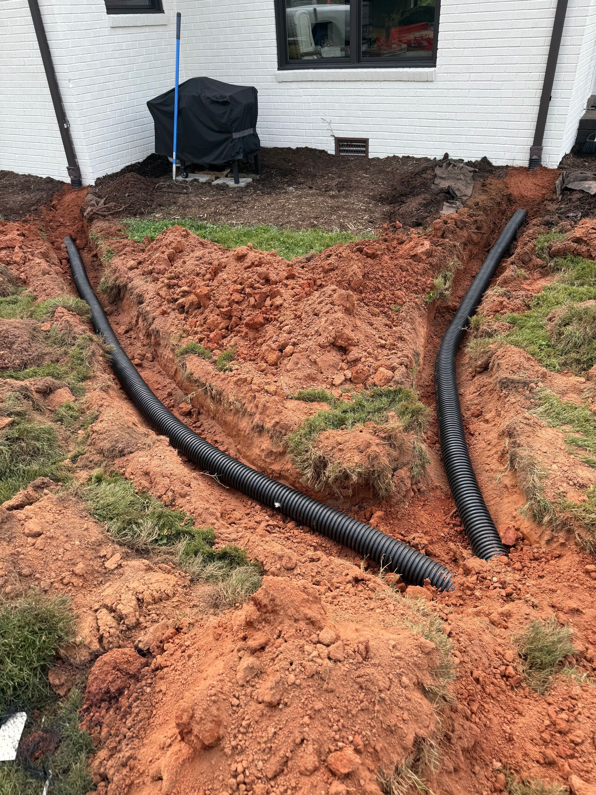 Two black corrugated drainage pipes are laid in trenches in red clay soil leading away from a house foundation.