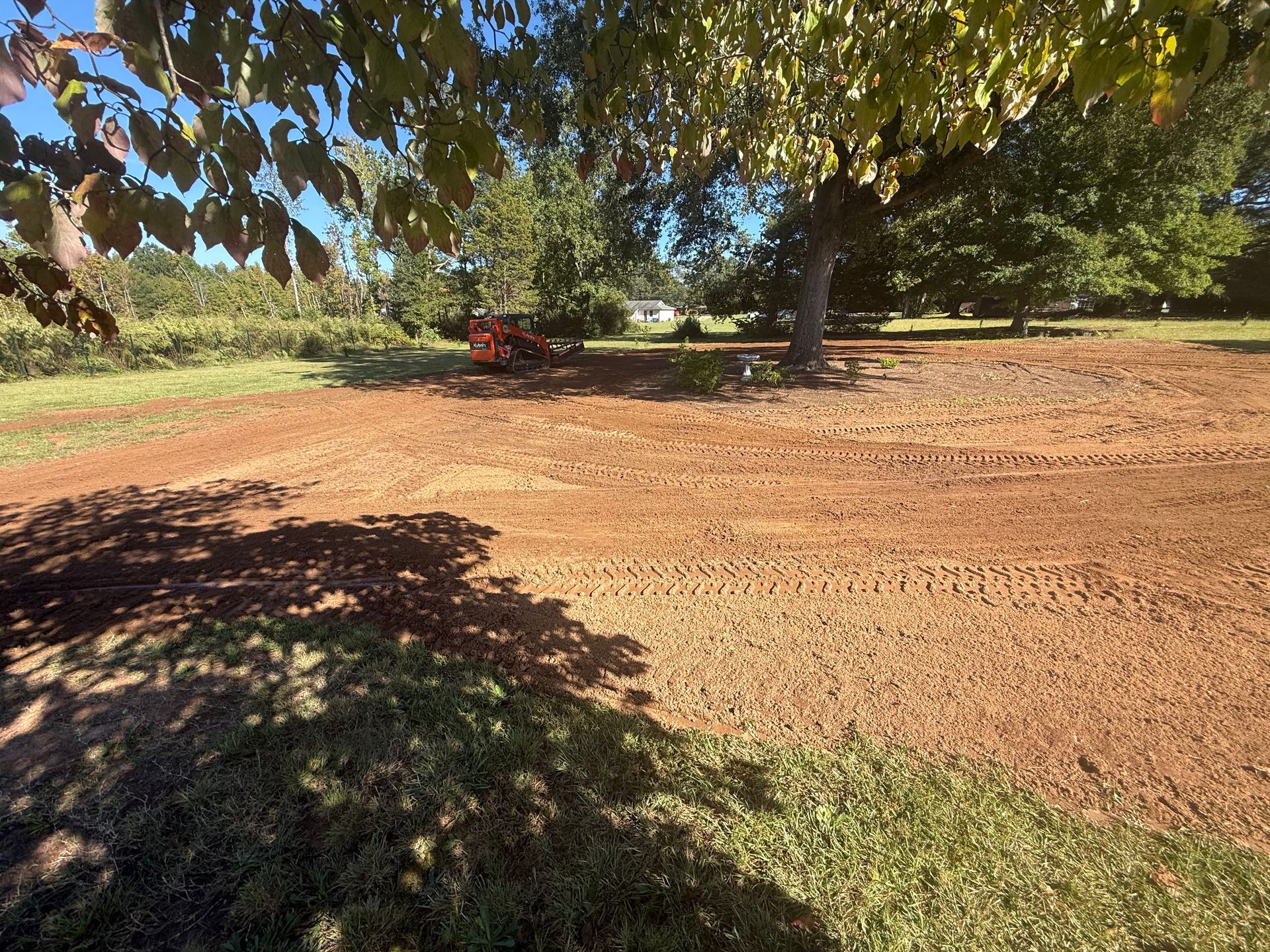 A red tractor works in a large, freshly tilled, reddish-brown soil field under trees on a sunny day.
