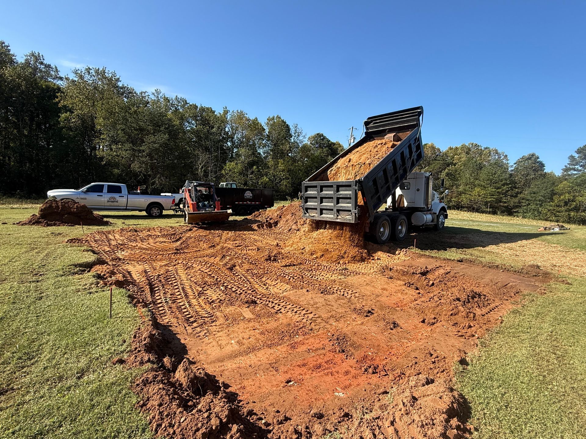 A dump truck pours soil into a muddy construction site in a grassy field with trees in the background.