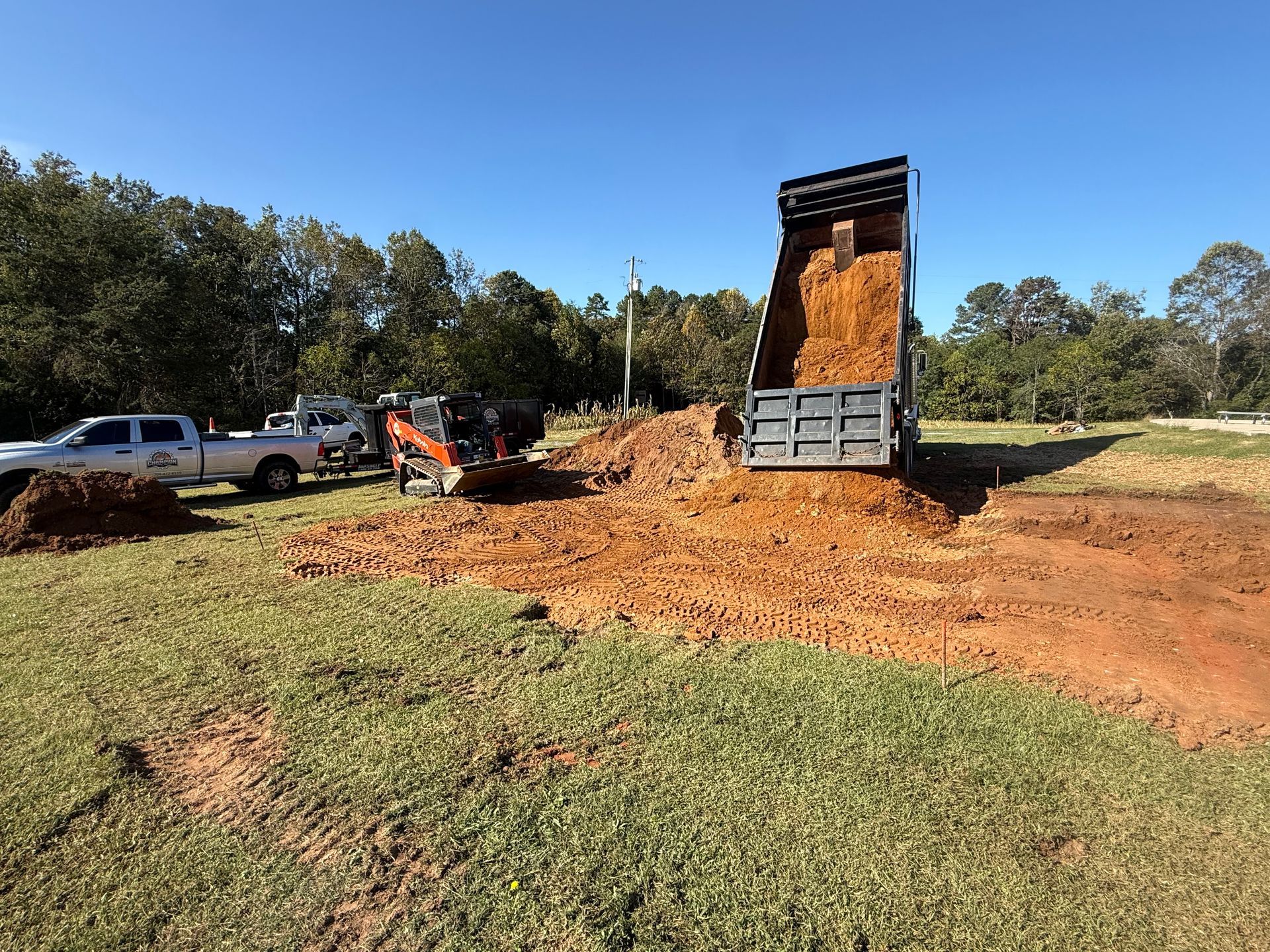 A dump truck tips a pile of red dirt onto a grassy site, with a pickup truck and small skid-steer loader nearby.