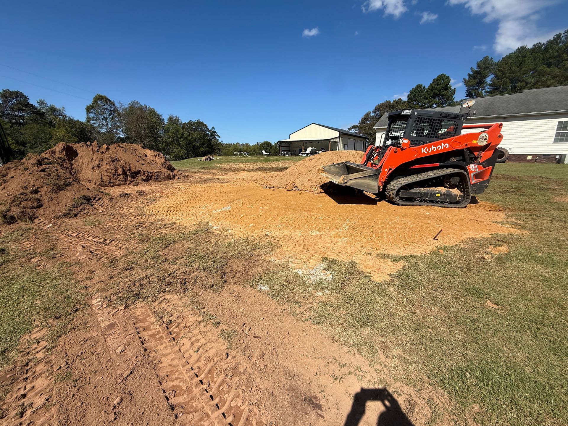 An orange skid steer loader levels dirt in a grassy residential yard on a sunny day.
