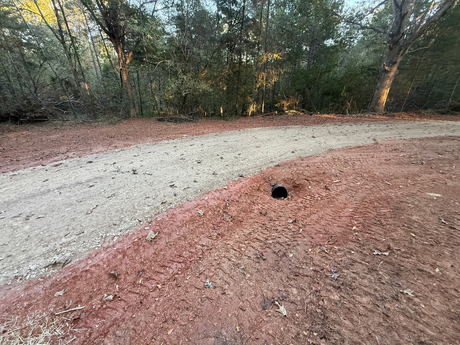 A black culvert pipe sits embedded in red dirt along the edge of a gravel driveway lined with trees.