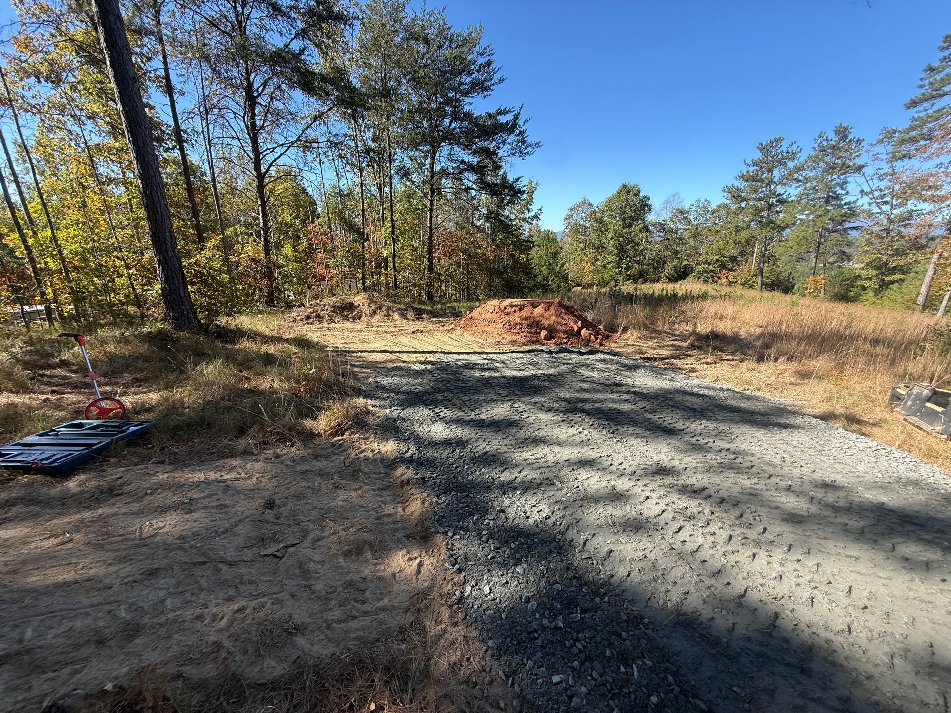 A gravel driveway leads through a sunny, wooded clearing toward a pile of dirt, with a mechanical tool on the left.