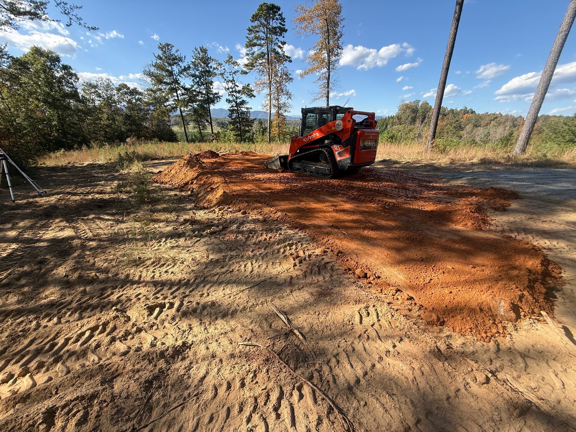 An orange skid-steer loader sits on a patch of freshly dug red clay dirt in a rural, sunny, wooded landscape.