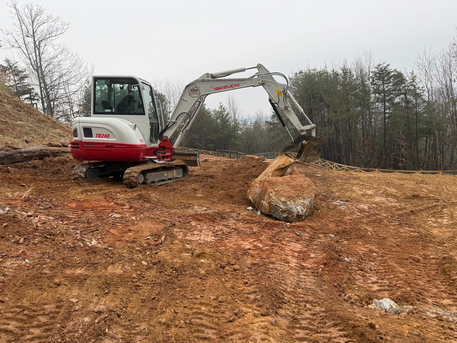 A white and red excavator moves a large rock on a dirt hillside against a backdrop of trees.