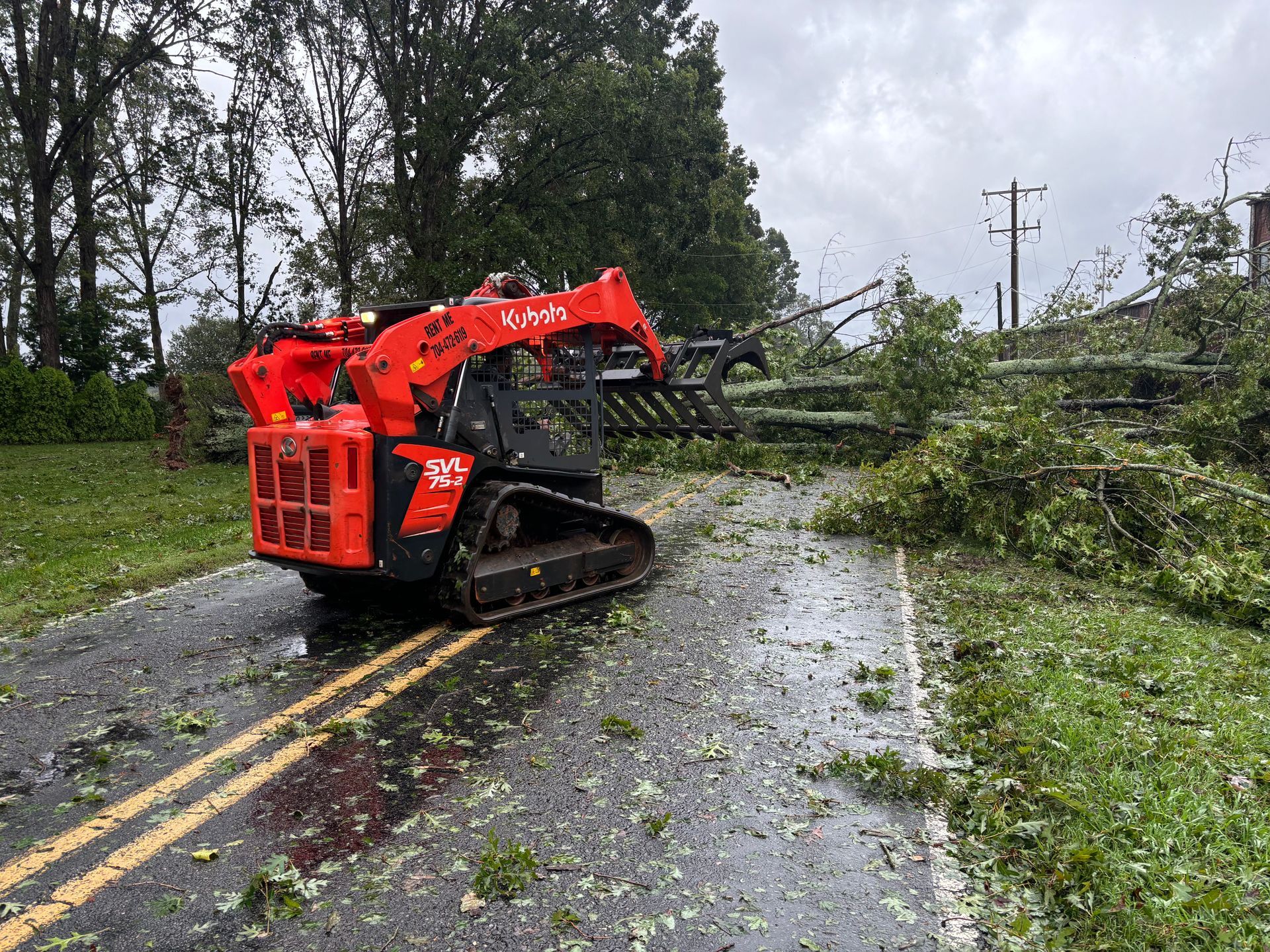 A bright orange skid steer with a grapple attachment clears fallen tree branches from a road after a storm.