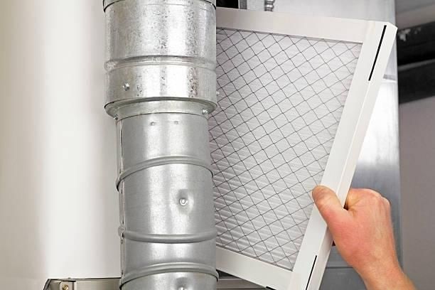 Hand removing a white furnace filter beside a metal air duct, showing dust buildup