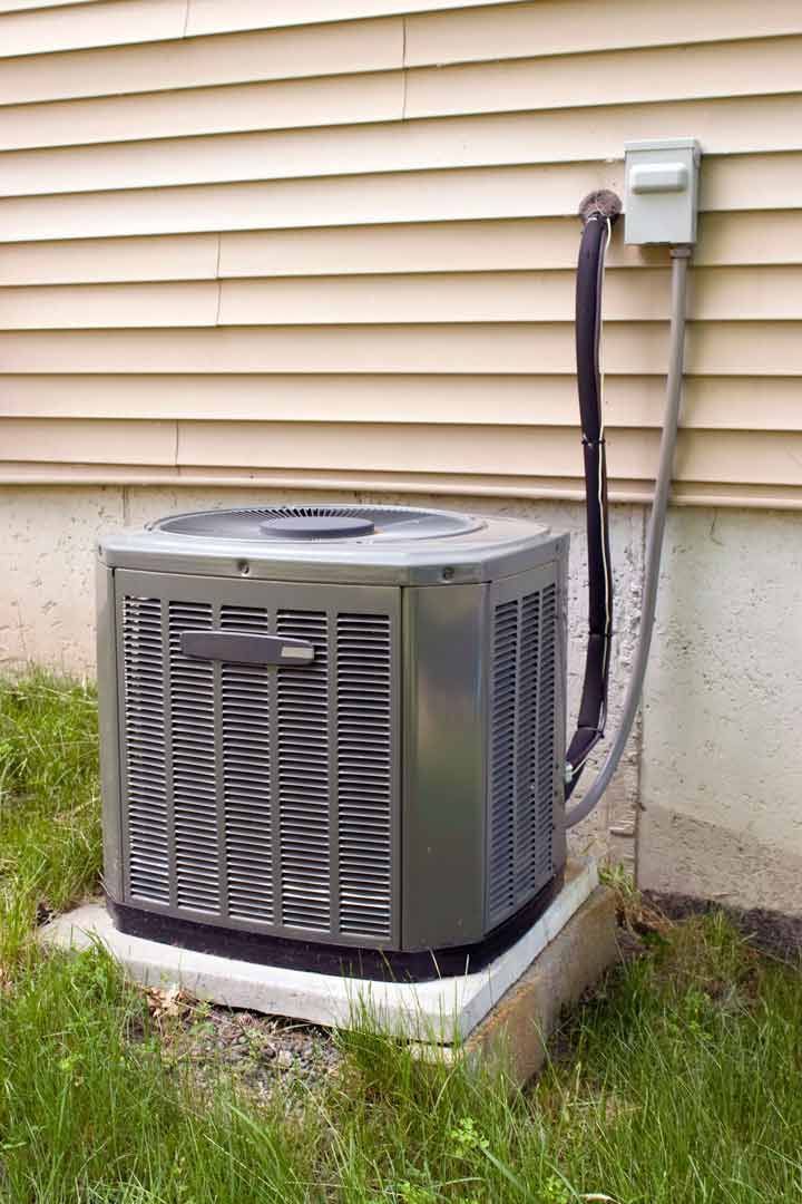 Outdoor air conditioner unit beside a house with wall-mounted electrical box and hoses along beige siding