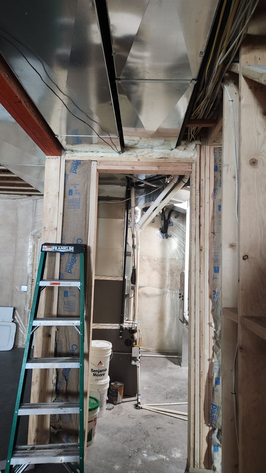 Narrow unfinished hallway with exposed ceiling pipes, a blue ladder, and construction materials.