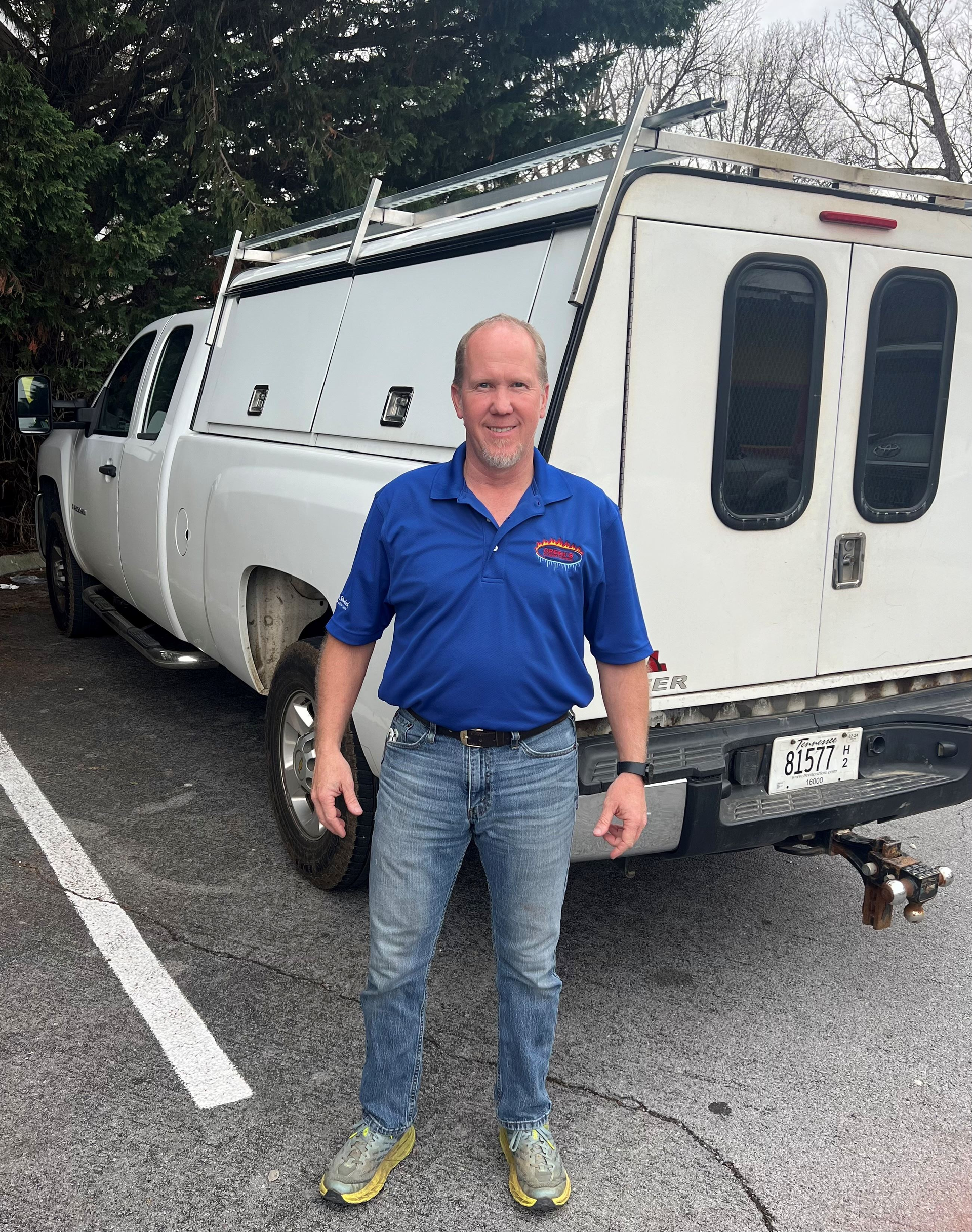 Man in blue shirt and jeans stands in front of a white truck with a camper shell.