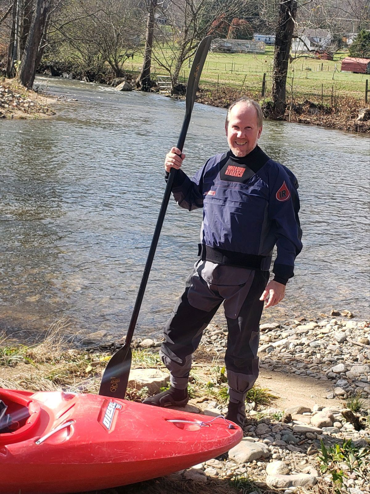 Man in dry suit with kayak paddle, standing by a red kayak near a river.