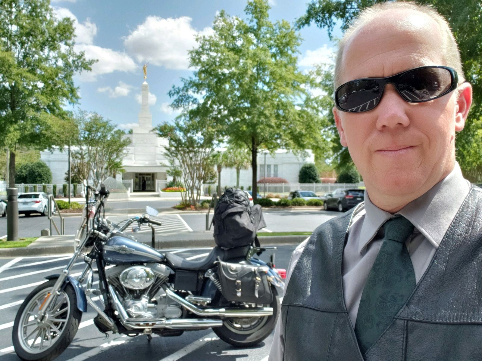 Man in sunglasses and vest by a parked Harley-Davidson motorcycle in front of a white church-like building.