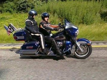 Two people on a blue Harley-Davidson motorcycle with US flags, riding on a road.